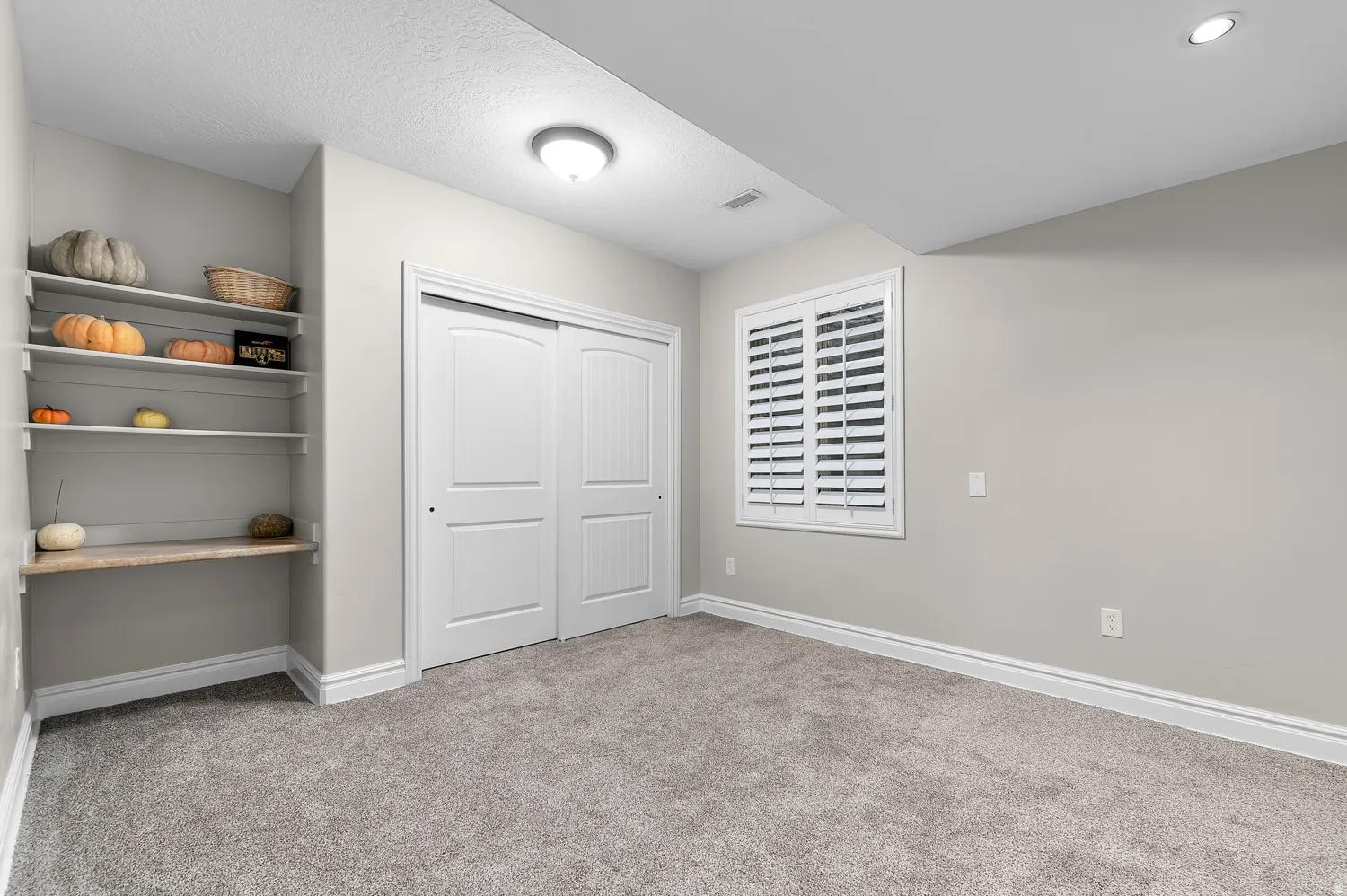 Carpeted interior space featuring a built-in shelving unit with a wood-finish desk surface, a dual-door closet, and a window with white plantation shutters