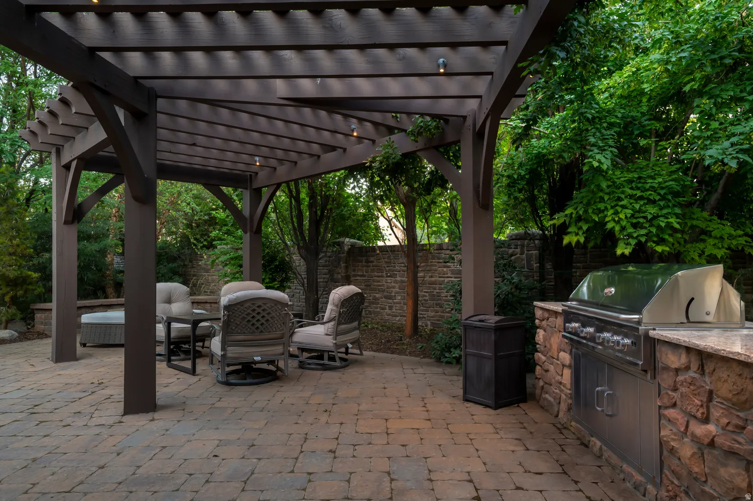 Expansive paver patio featuring a dark-stained wood pergola, integrated outdoor grill, stone-faced outdoor kitchen counter, and surrounding mature trees