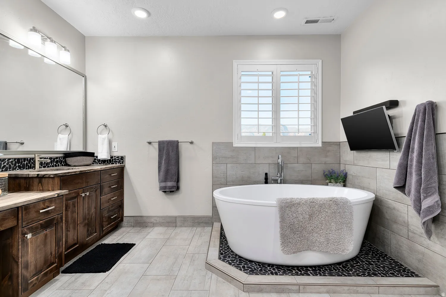 Freestanding soaking tub with pebble tile surround, accompanied by wood cabinetry with stone countertops, a large vanity mirror, and wall-mounted television