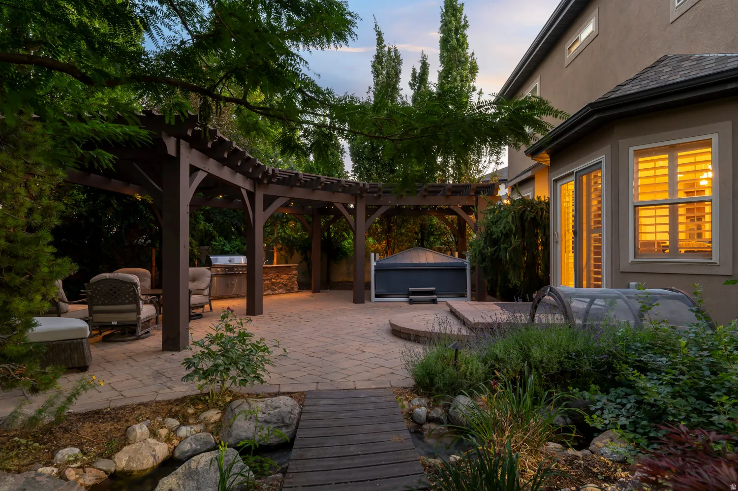 Expansive paver patio featuring a multi-section wood pergola, built-in outdoor kitchen with grill, and an integrated hot tub