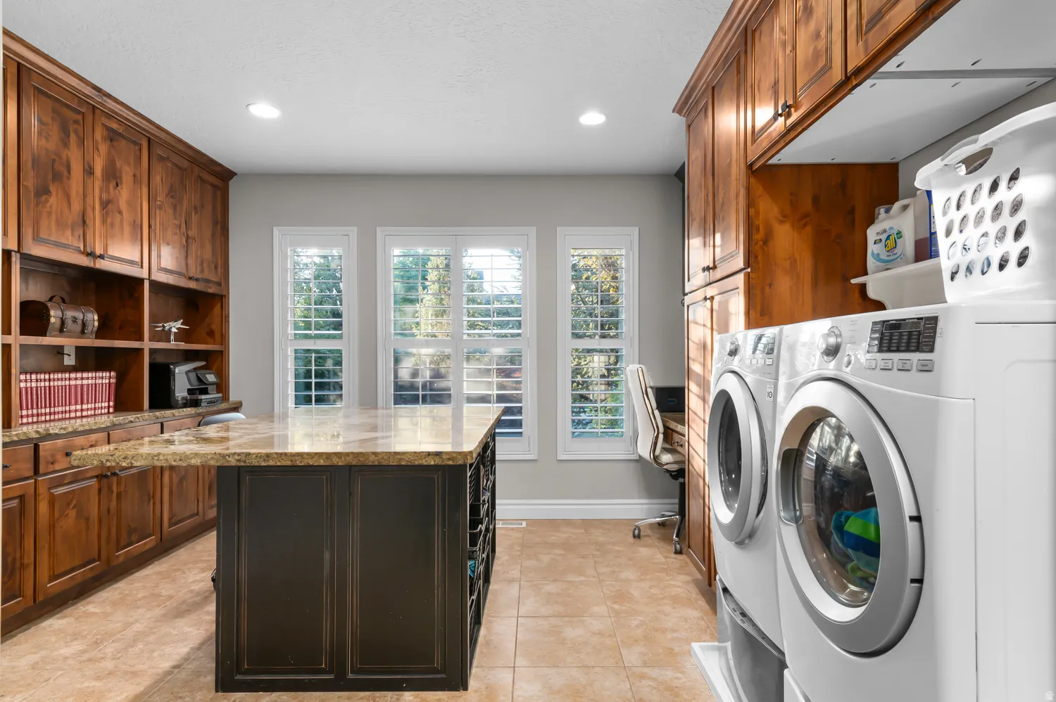 Spacious laundry room featuring a large central island with a stone countertop, extensive wood cabinetry, and three windows with plantation shutters