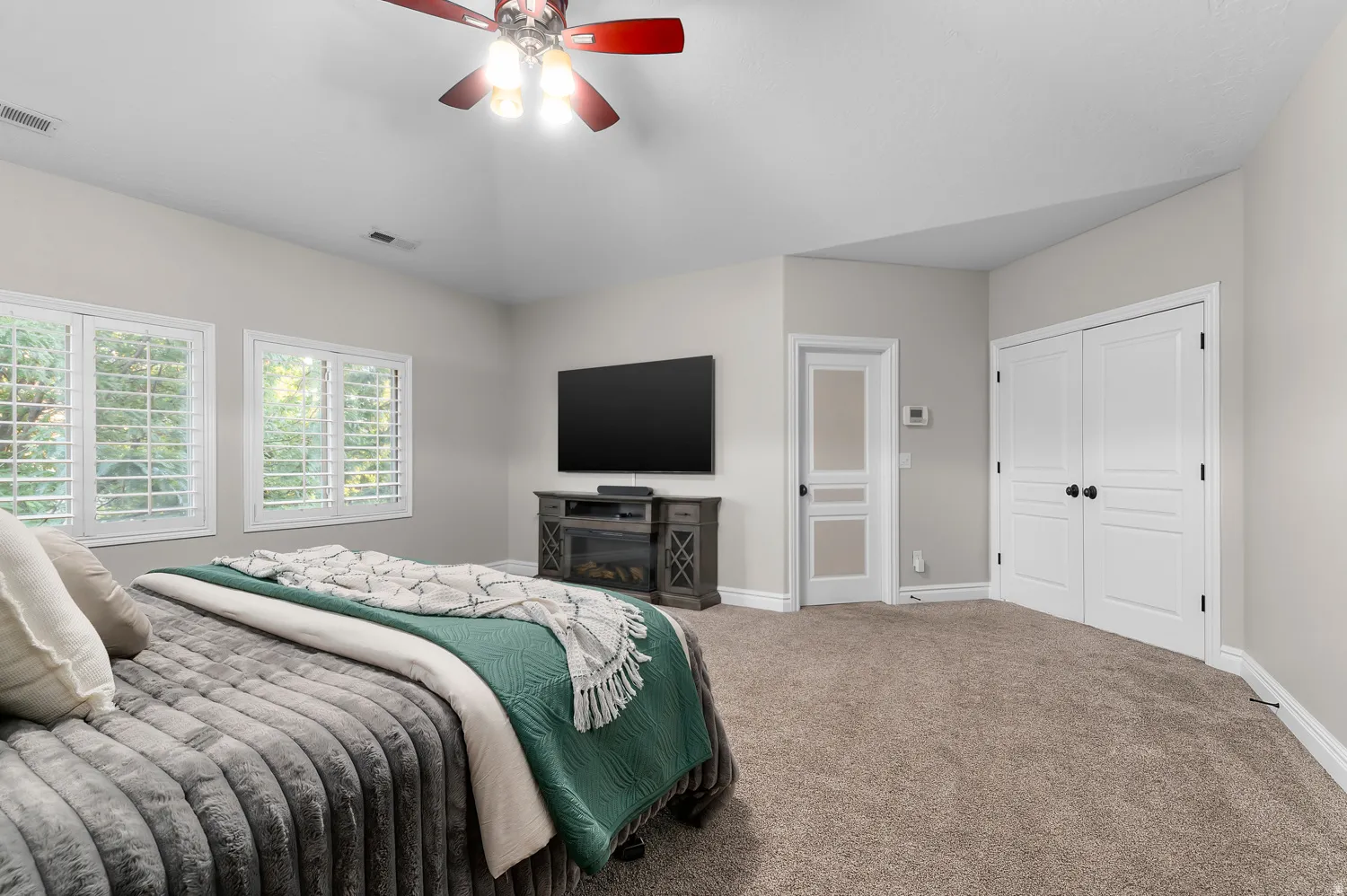 Carpeted room featuring a ceiling fan, dual windows with white plantation shutters, and a wall-mounted television above a wood-finish media console