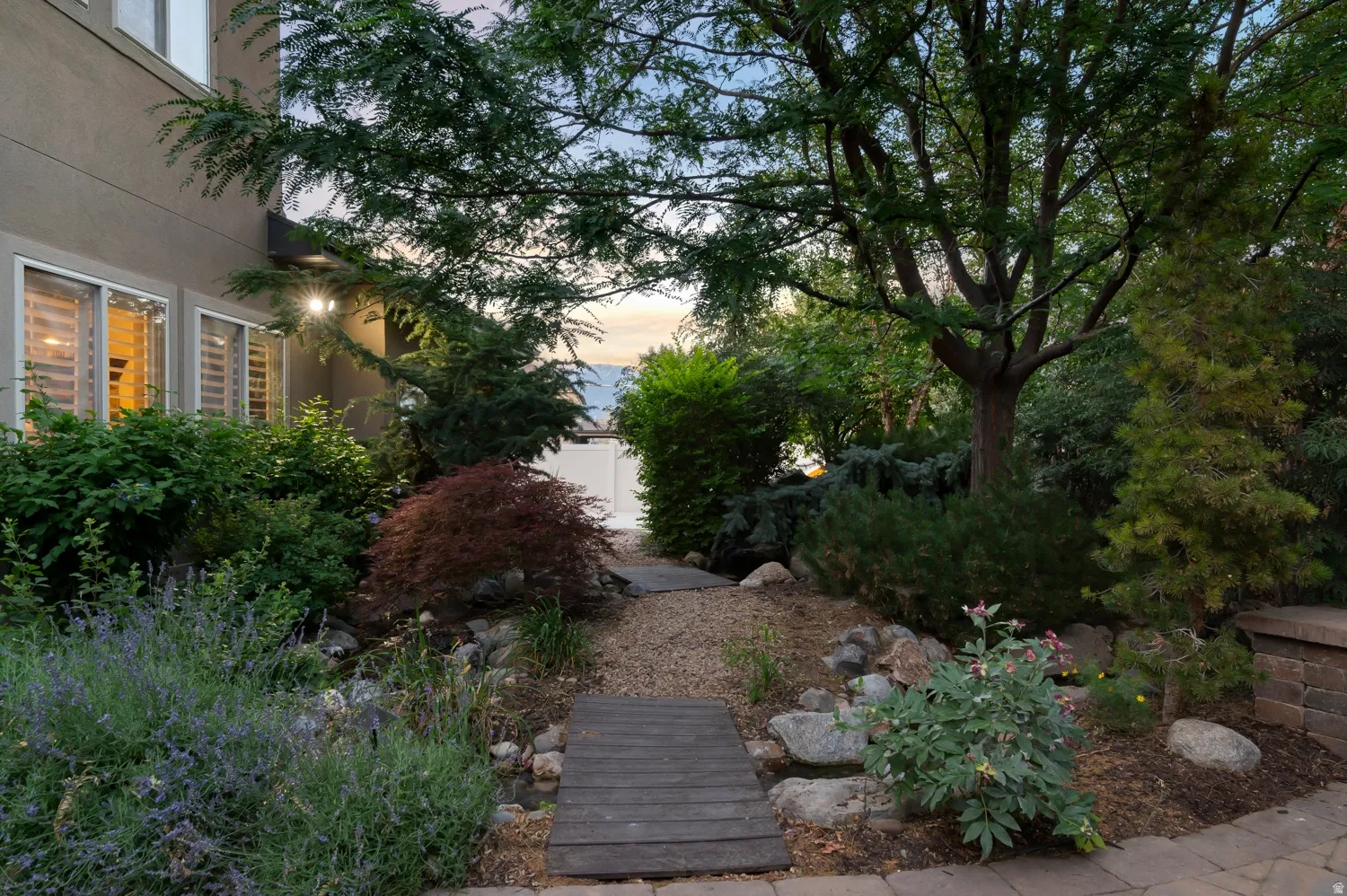 Landscaped exterior featuring a gravel pathway with wooden stepping stones, mature trees, diverse shrubbery, and decorative rockwork