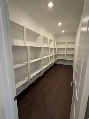 Expansive walk-in pantry featuring abundant white shelving, dark wood-finish flooring, recessed lighting, and white walls