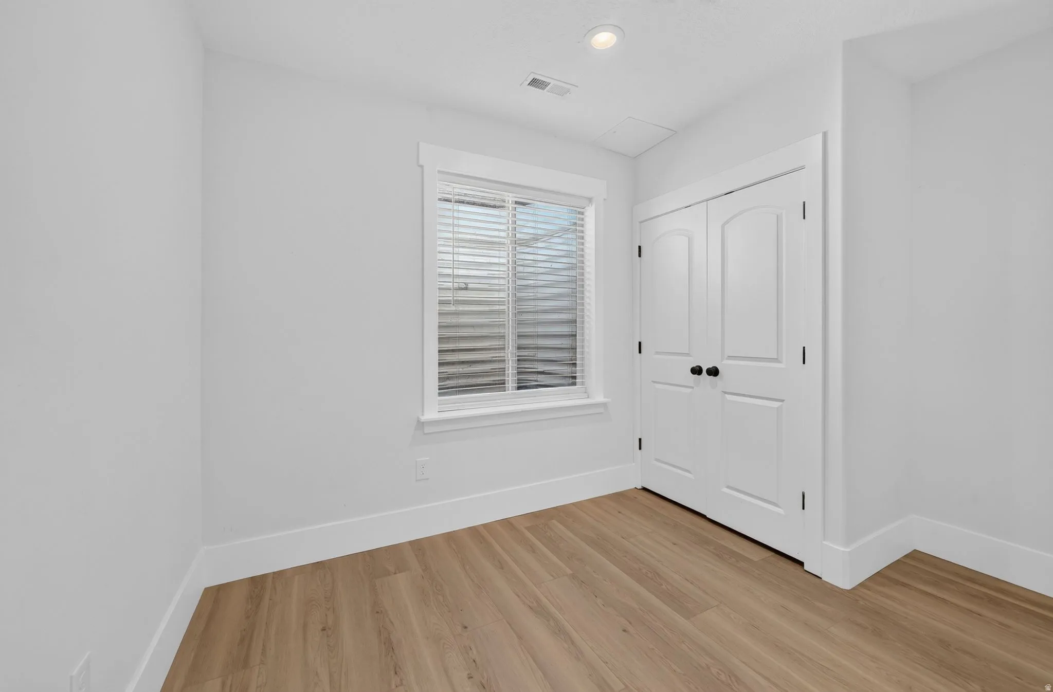 Bright room featuring wood-finish flooring, a double-door closet with black hardware, a window with horizontal blinds, recessed lighting, and white trim throughout