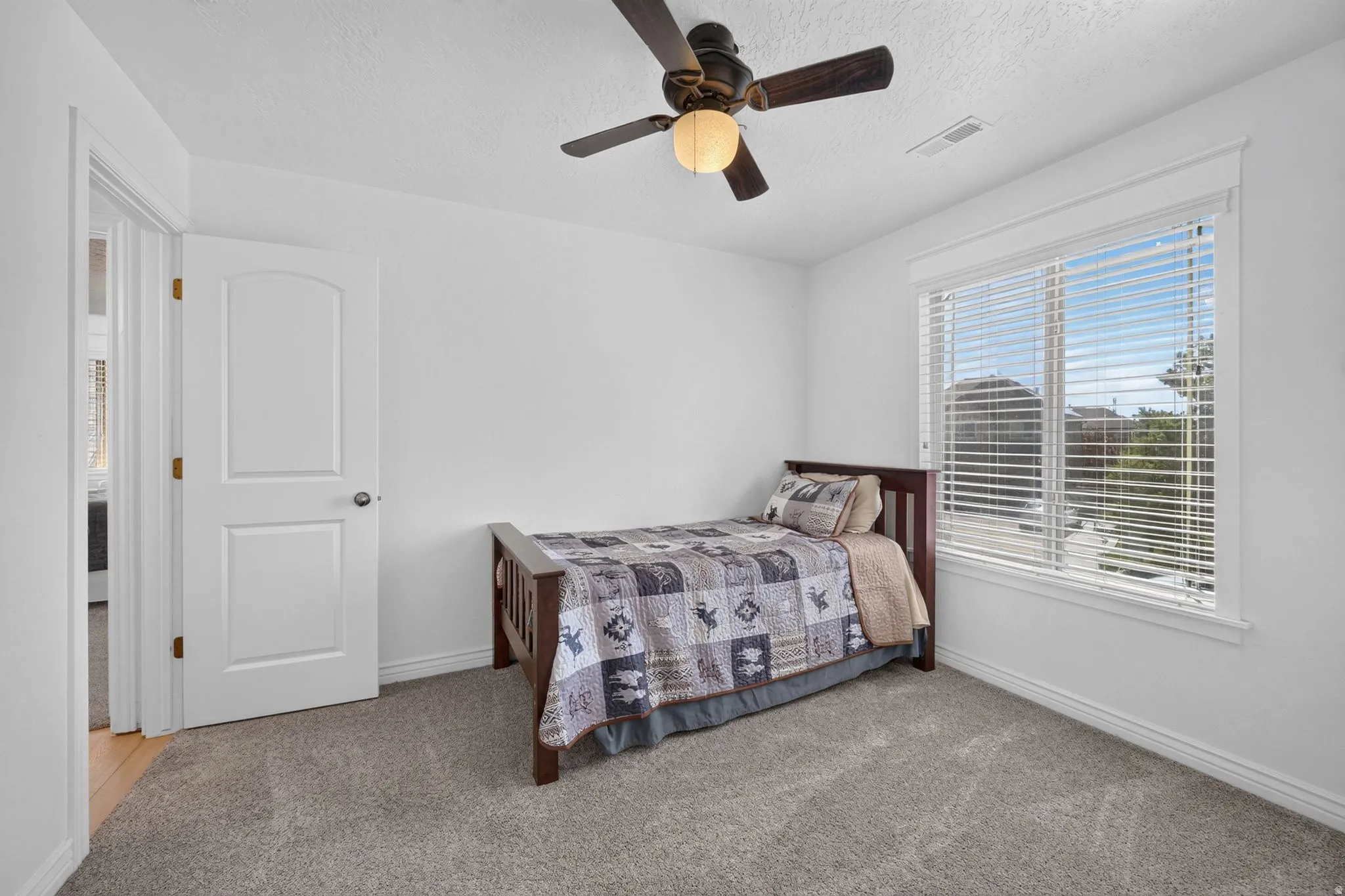 Bright room featuring a large window with blinds, a ceiling fan with integrated lighting, white walls, and gray carpet flooring