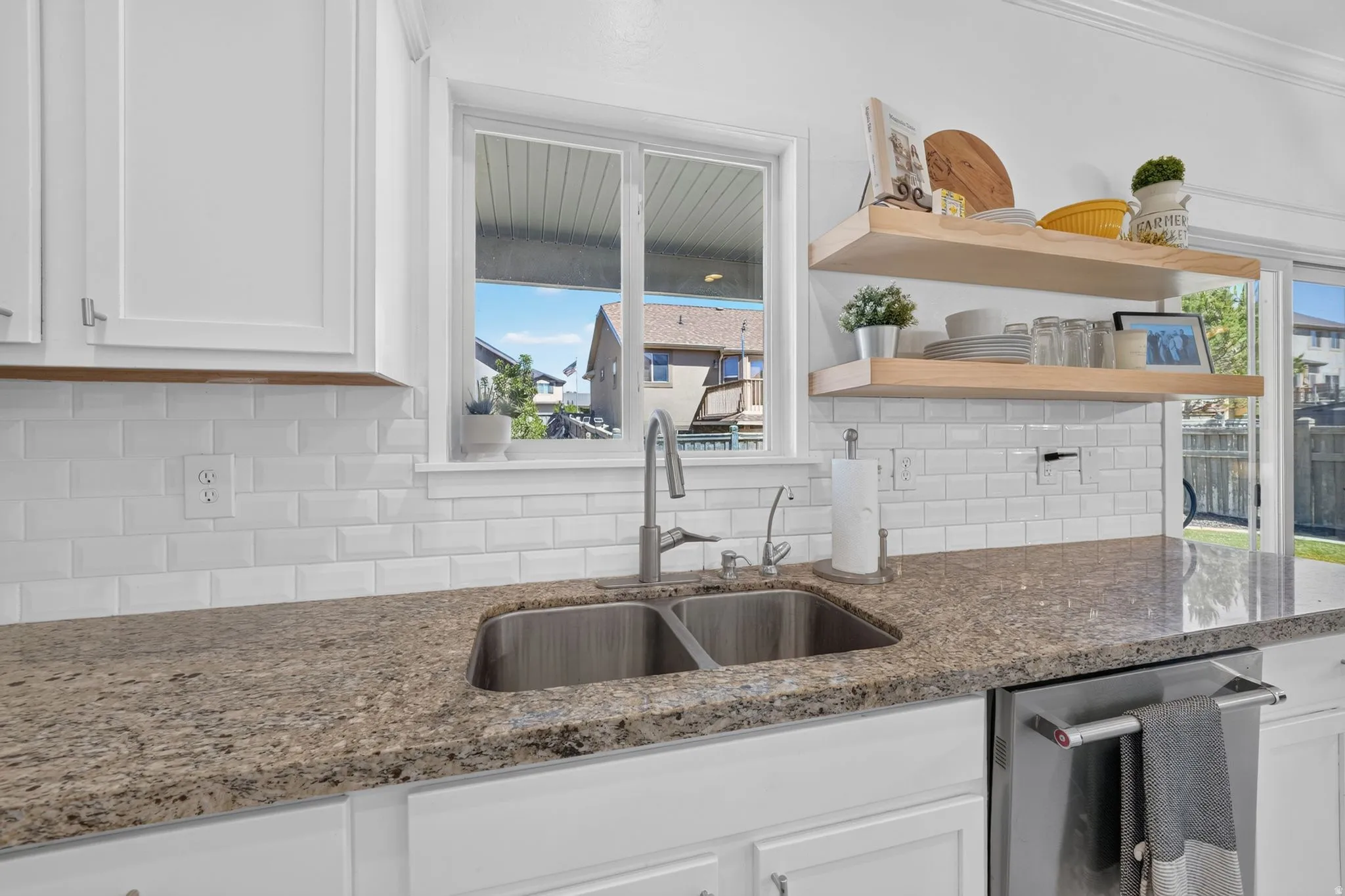 Kitchen featuring a double basin stainless steel sink, solid surface countertops, white subway tile backsplash, white cabinetry, and open wood shelving