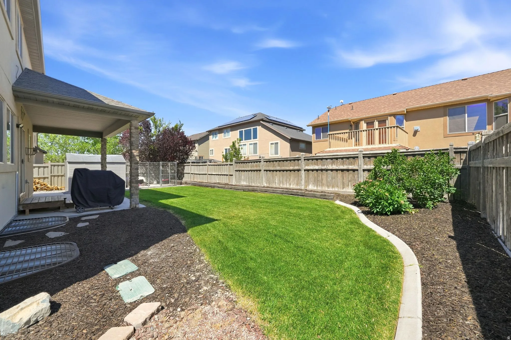 Expansive backyard featuring a lush green lawn, mature landscaping, wood fencing, a covered patio, and stepping stone pathways