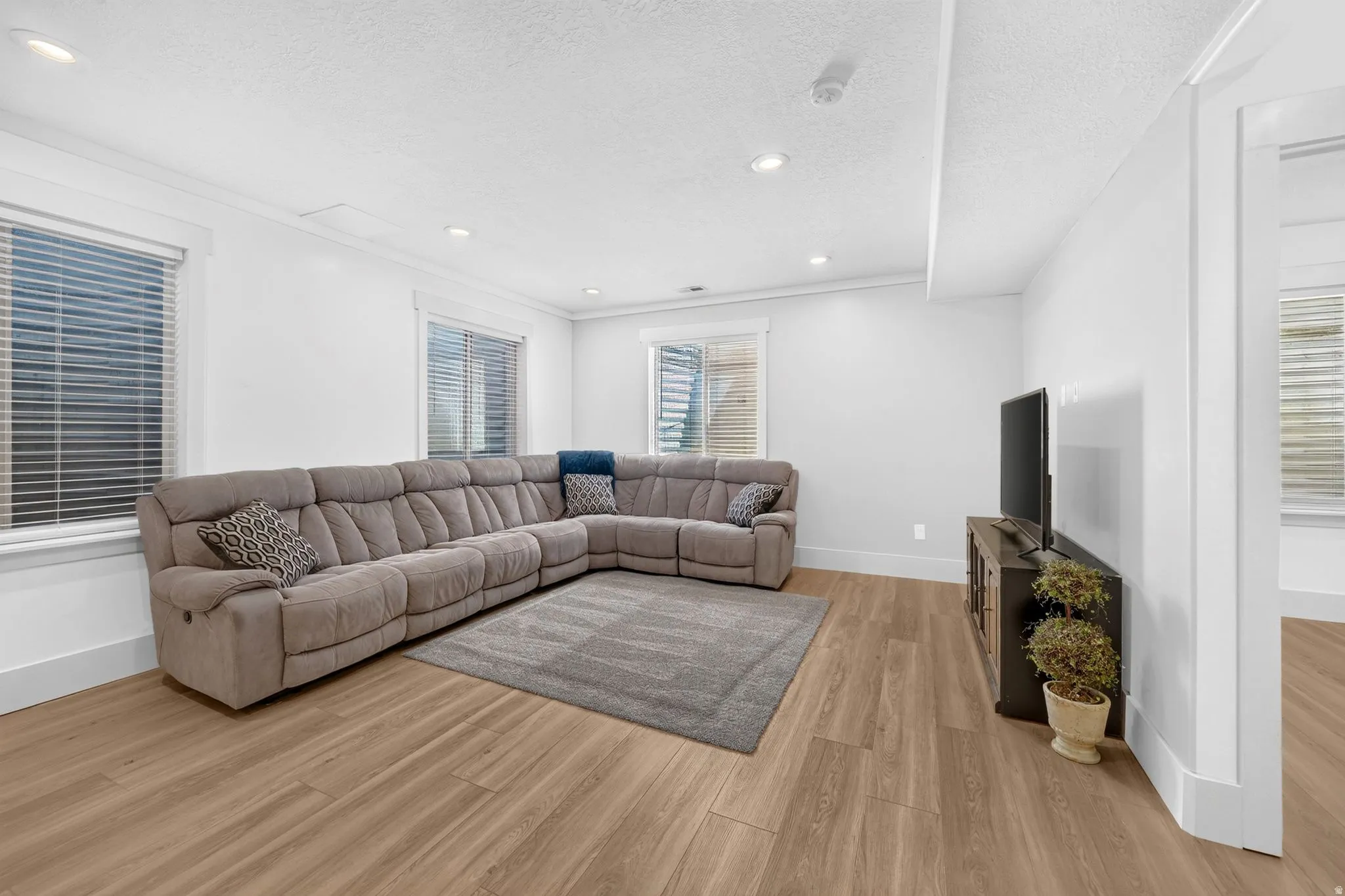 Spacious living area featuring light wood-finish flooring, recessed lighting, and multiple windows with horizontal blinds