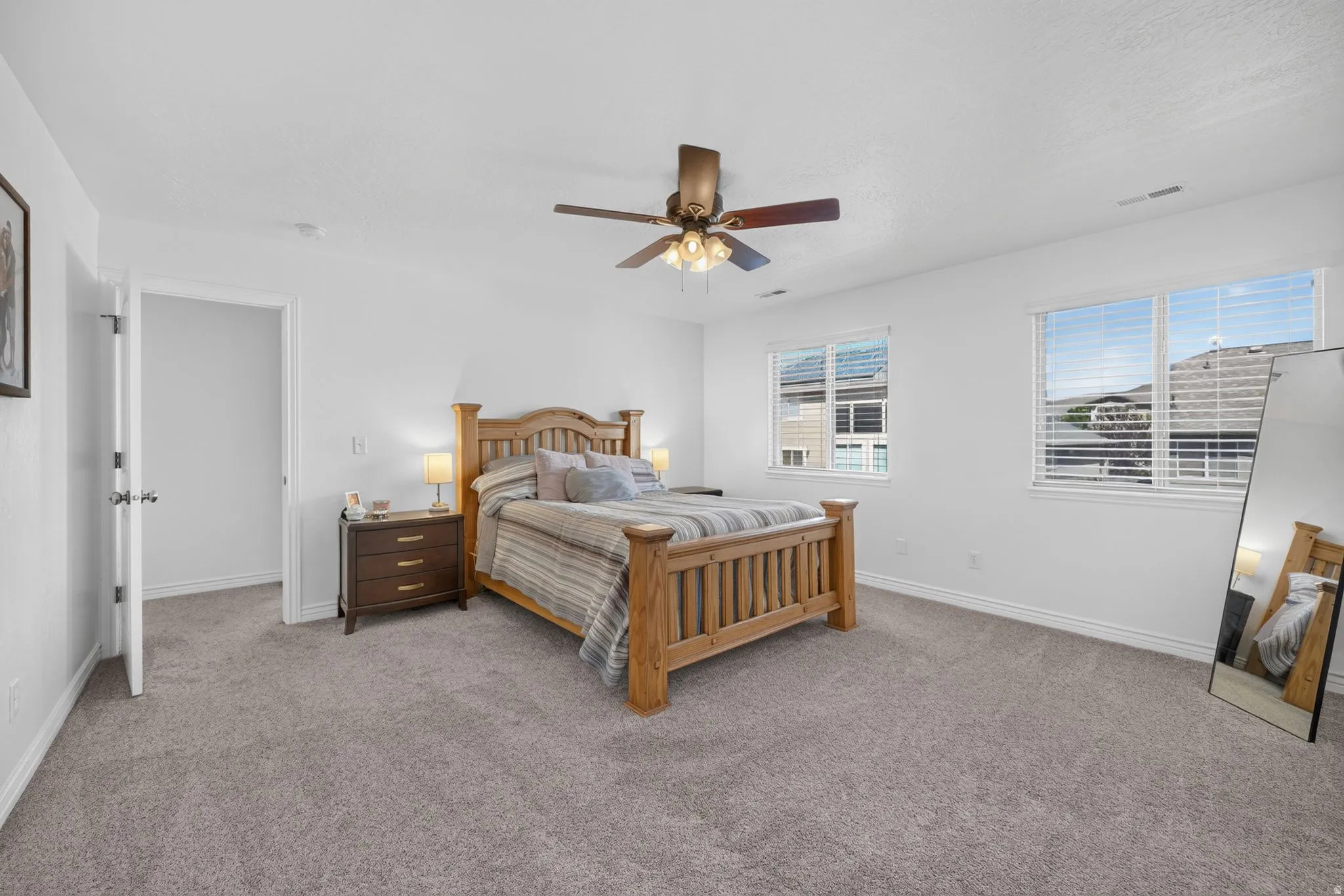 Spacious bedroom featuring light gray carpeting, crisp white walls, two large windows with blinds, a ceiling fan with integrated lighting, and a white interior door