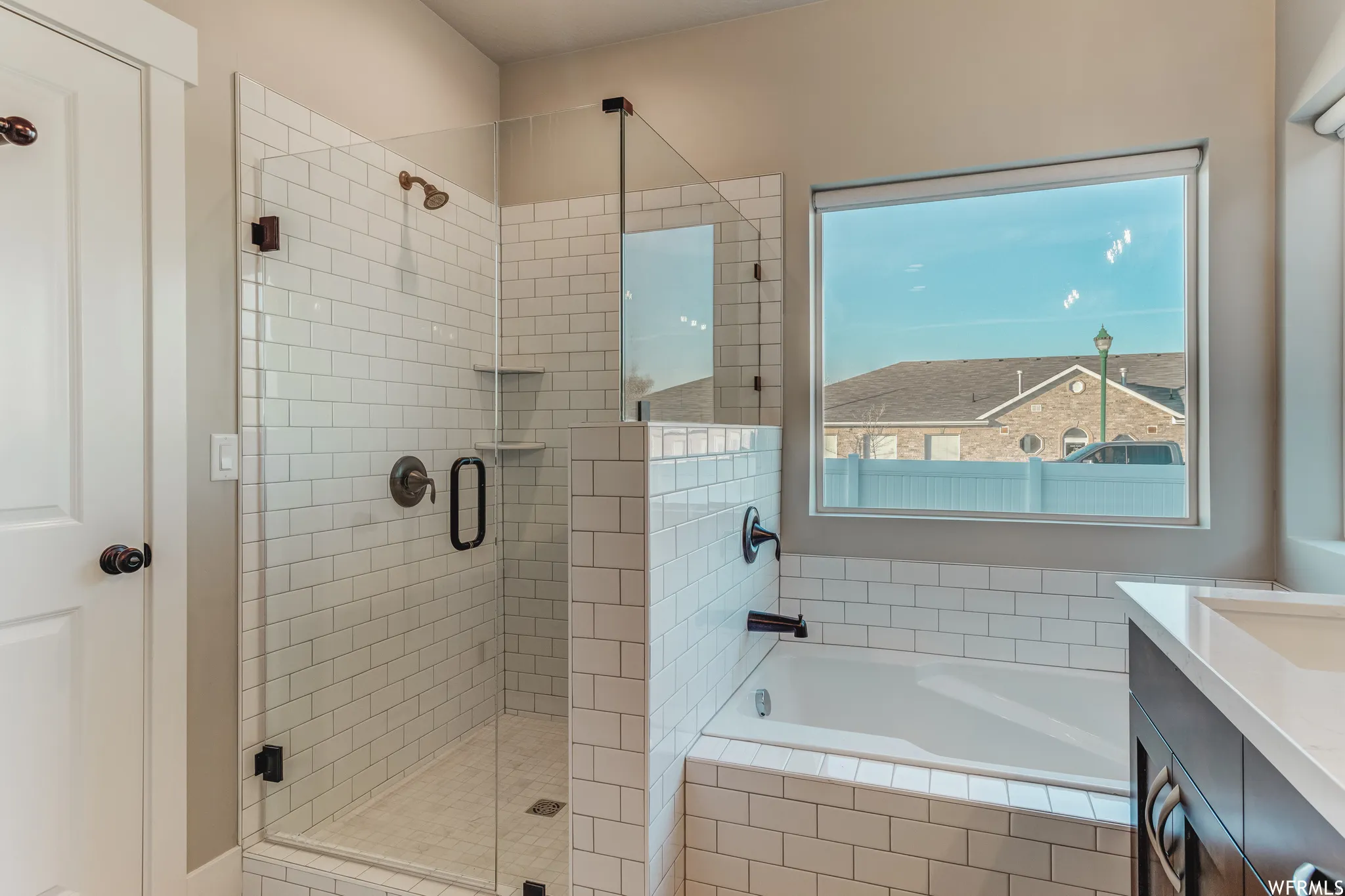 Spa-inspired primary bathroom featuring a frameless glass-enclosed walk-in shower with subway tile surround, a soaking tub positioned beneath a large window with natural light, dual vanity with quartz countertop, and matte black hardware and fixtures