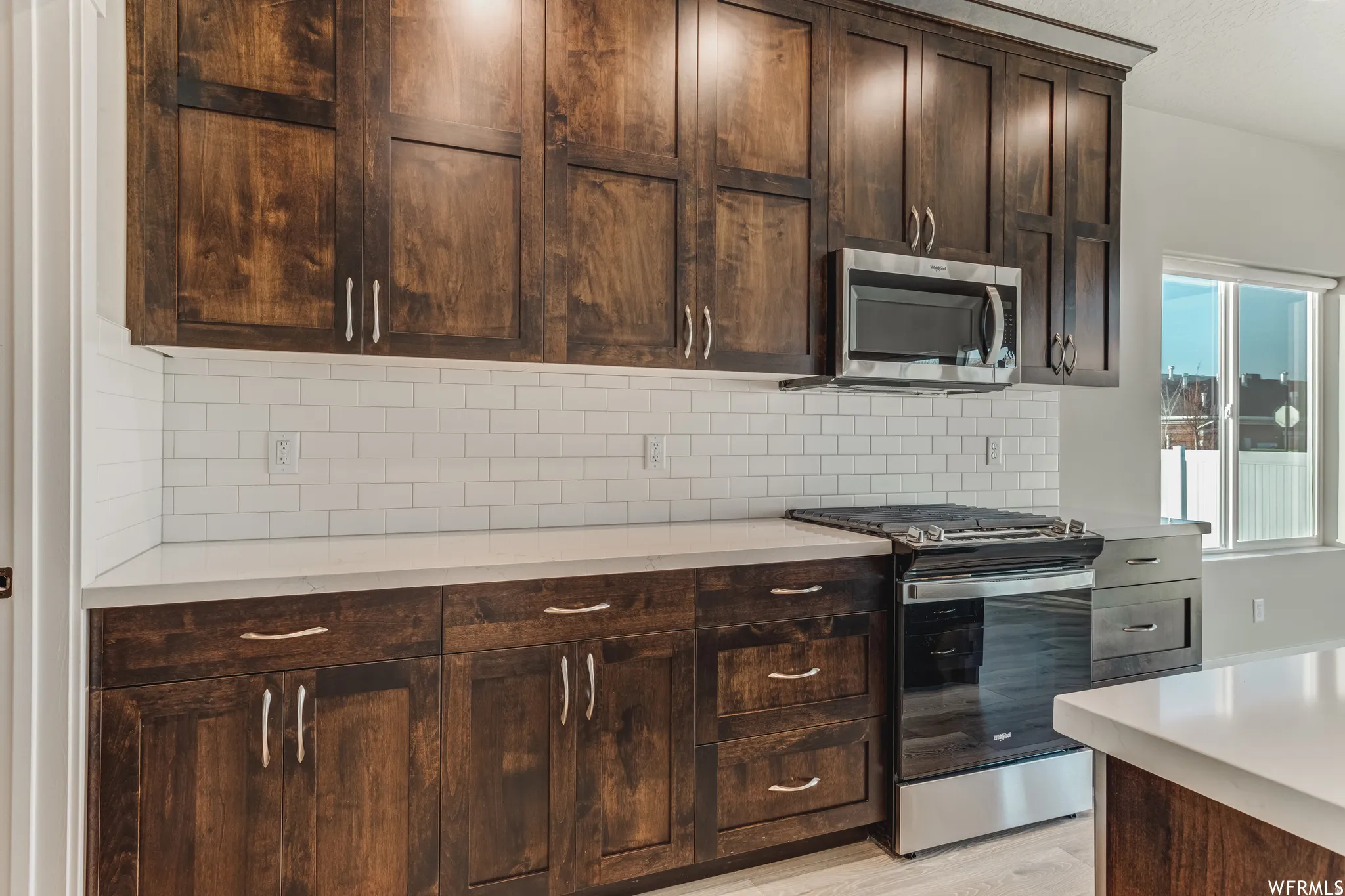 Dark wood-finish cabinetry with silver hardware, white subway tile backsplash, white countertops, stainless steel microwave, and stainless steel range