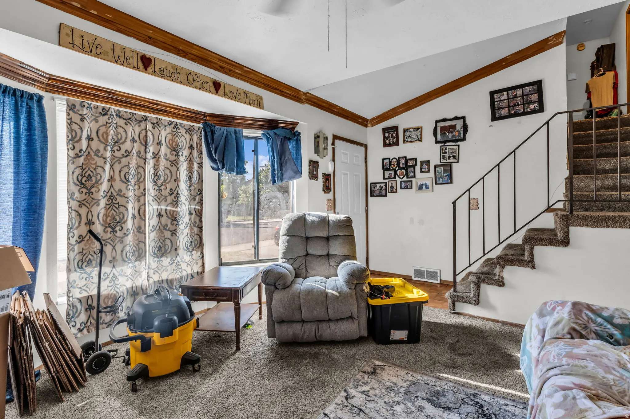 Spacious living area featuring a large bay window, neutral carpeting, white walls, and a staircase with a black metal railing