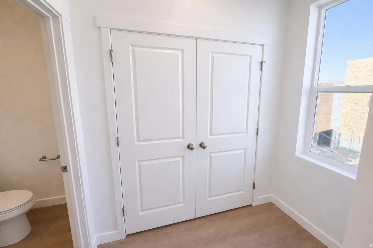 Interior space featuring a double-door closet with paneling and brushed nickel hardware, a single-hung window with white trim, light-colored flooring, and a neutral wall palette