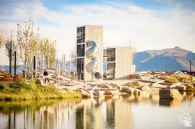 Waterfront recreational area featuring an architectural play structure with a spiral slide, natural wood climbing elements, and surrounding mature trees, all against a backdrop of distant mountains