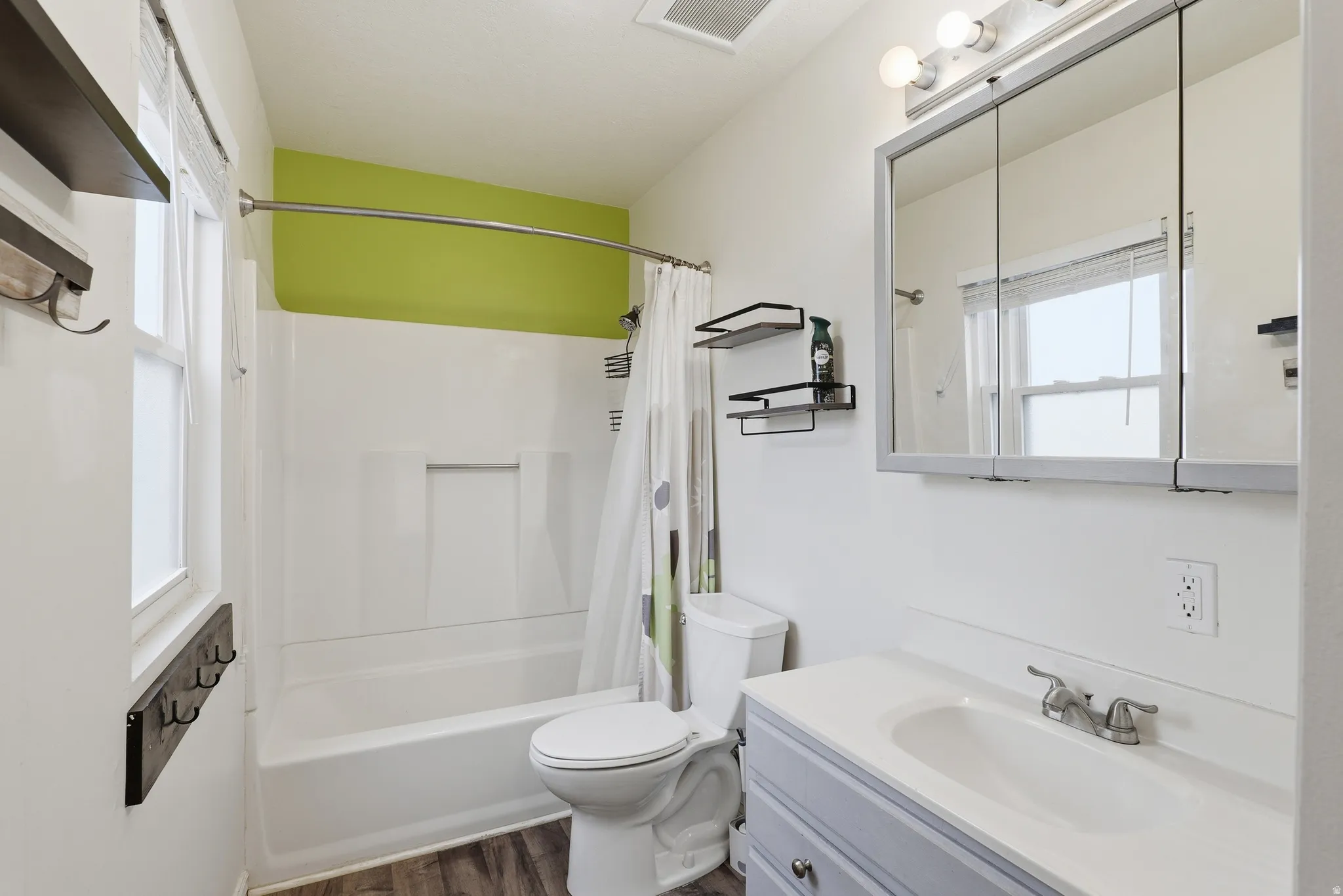 Bathroom featuring a white bathtub and shower surround, a light blue vanity with integrated sink, a mirrored medicine cabinet, wood-finish flooring, and a window