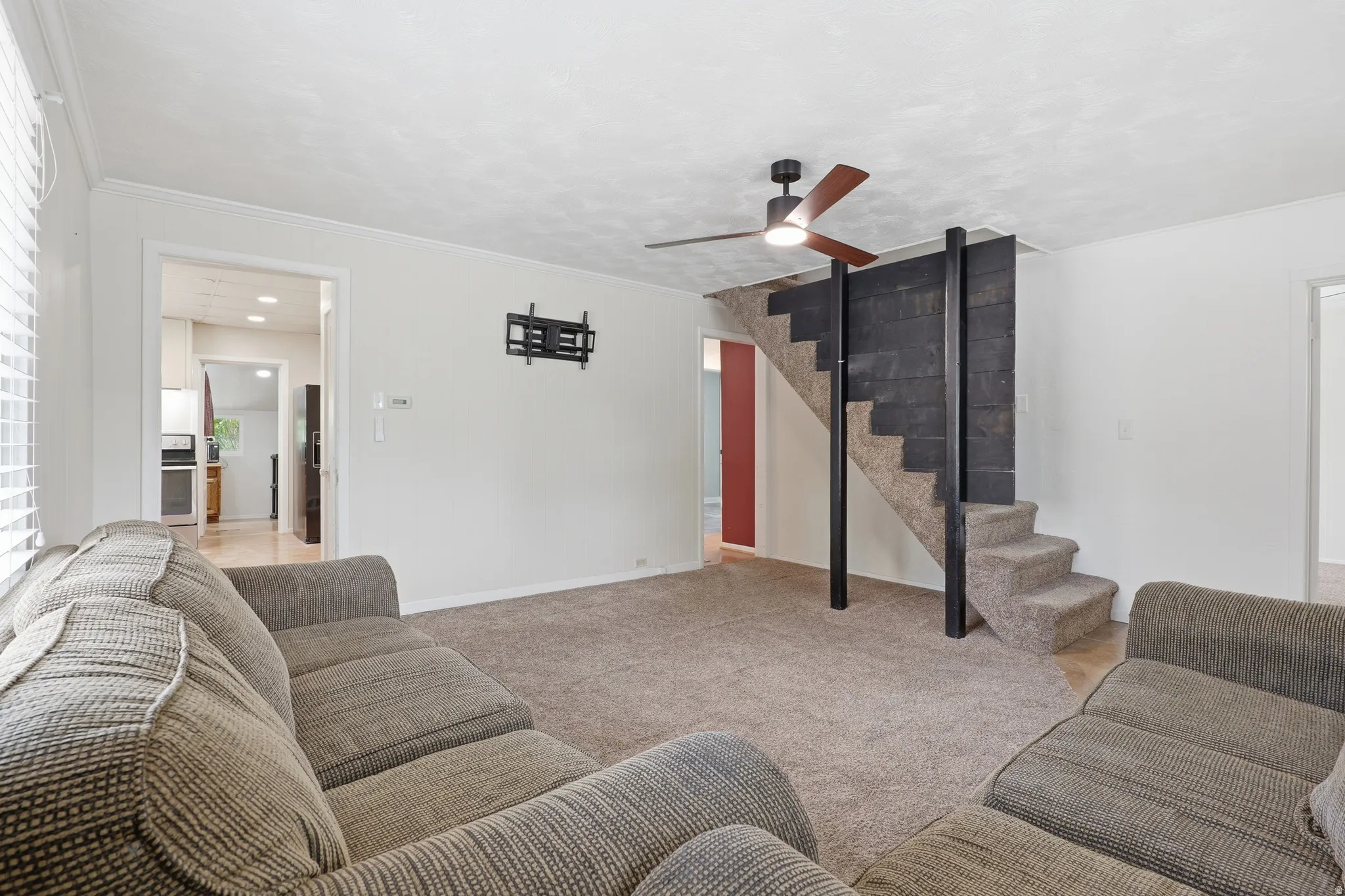 Carpeted living area featuring a modern ceiling fan with light, a textured accent wall bordering a carpeted staircase, and an open doorway leading to a kitchen with wood-finish flooring