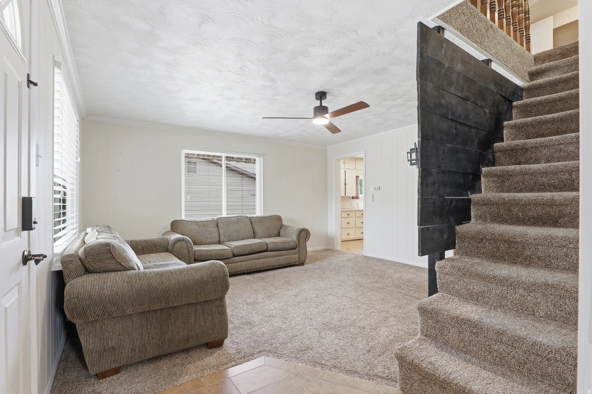 Carpeted living area featuring a ceiling fan, a window with blinds, and a built-in wall sconce, with a dark wood-finish partition framing the carpeted staircase