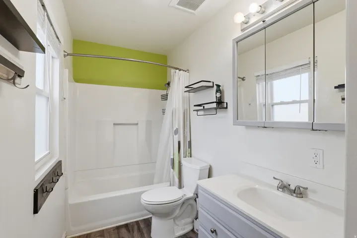Bathroom featuring a white bathtub and shower surround, a light blue vanity with integrated sink, a mirrored medicine cabinet, wood-finish flooring, and a window