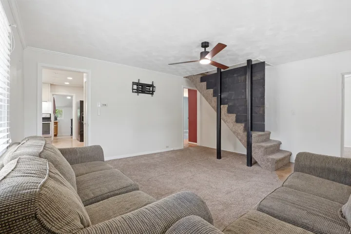 Carpeted living area featuring a modern ceiling fan with light, a textured accent wall bordering a carpeted staircase, and an open doorway leading to a kitchen with wood-finish flooring
