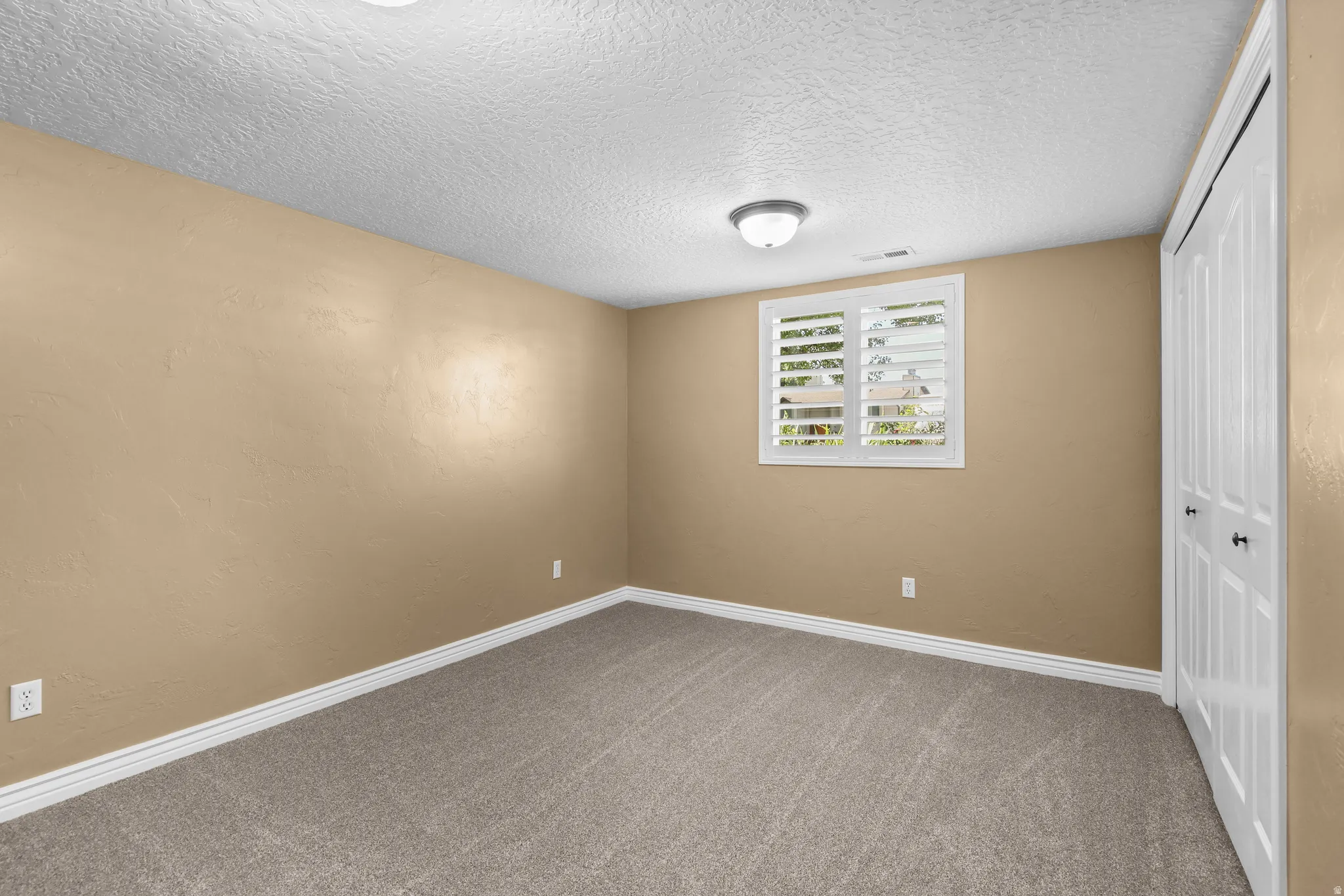 Neutral-toned carpeting in a spacious room featuring a window with white plantation shutters, a surface-mounted ceiling light fixture, and a white bi-fold closet door with black hardware