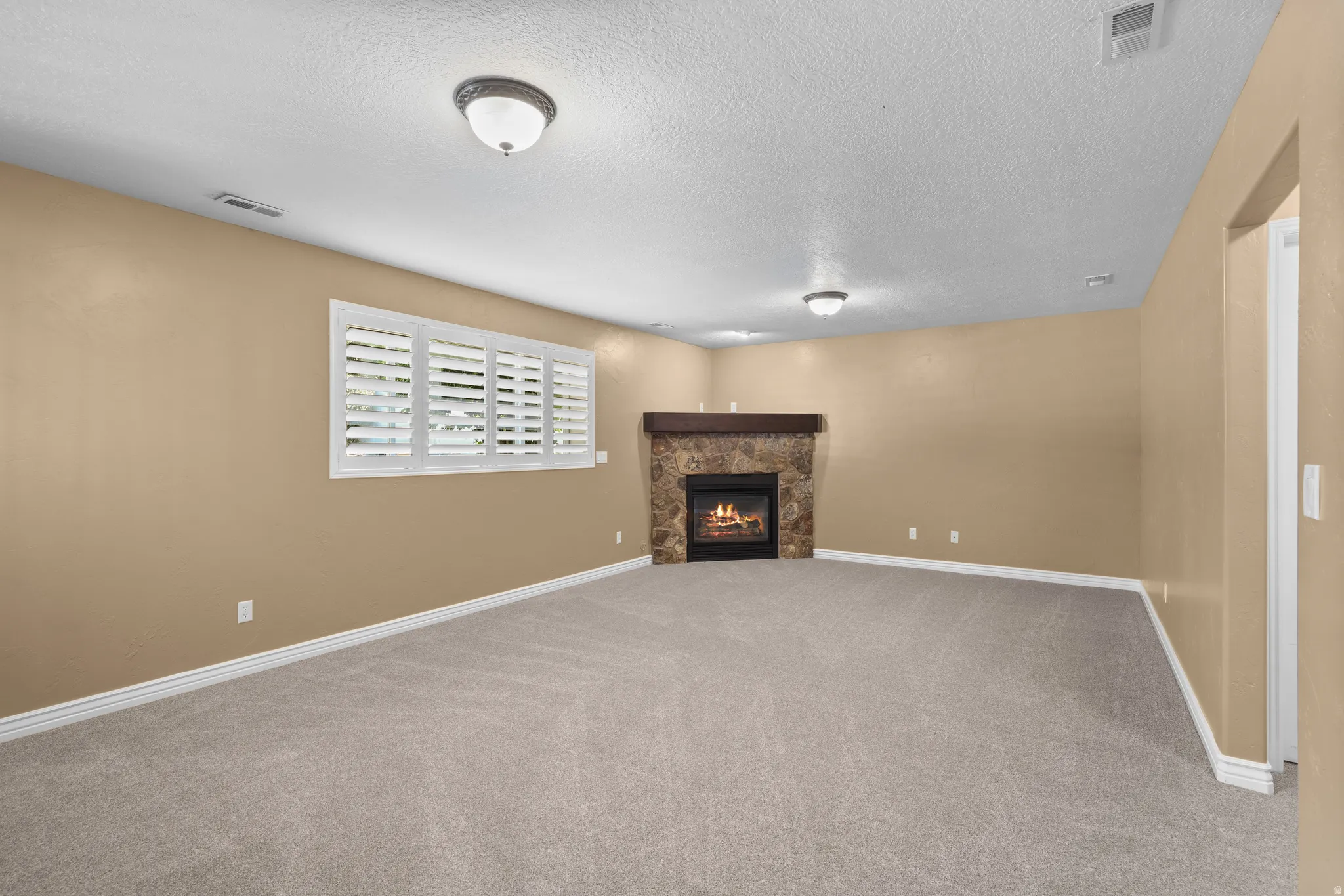 Spacious interior room featuring a stone fireplace with a wood-finish mantel, neutral carpeting, and a window with plantation shutters