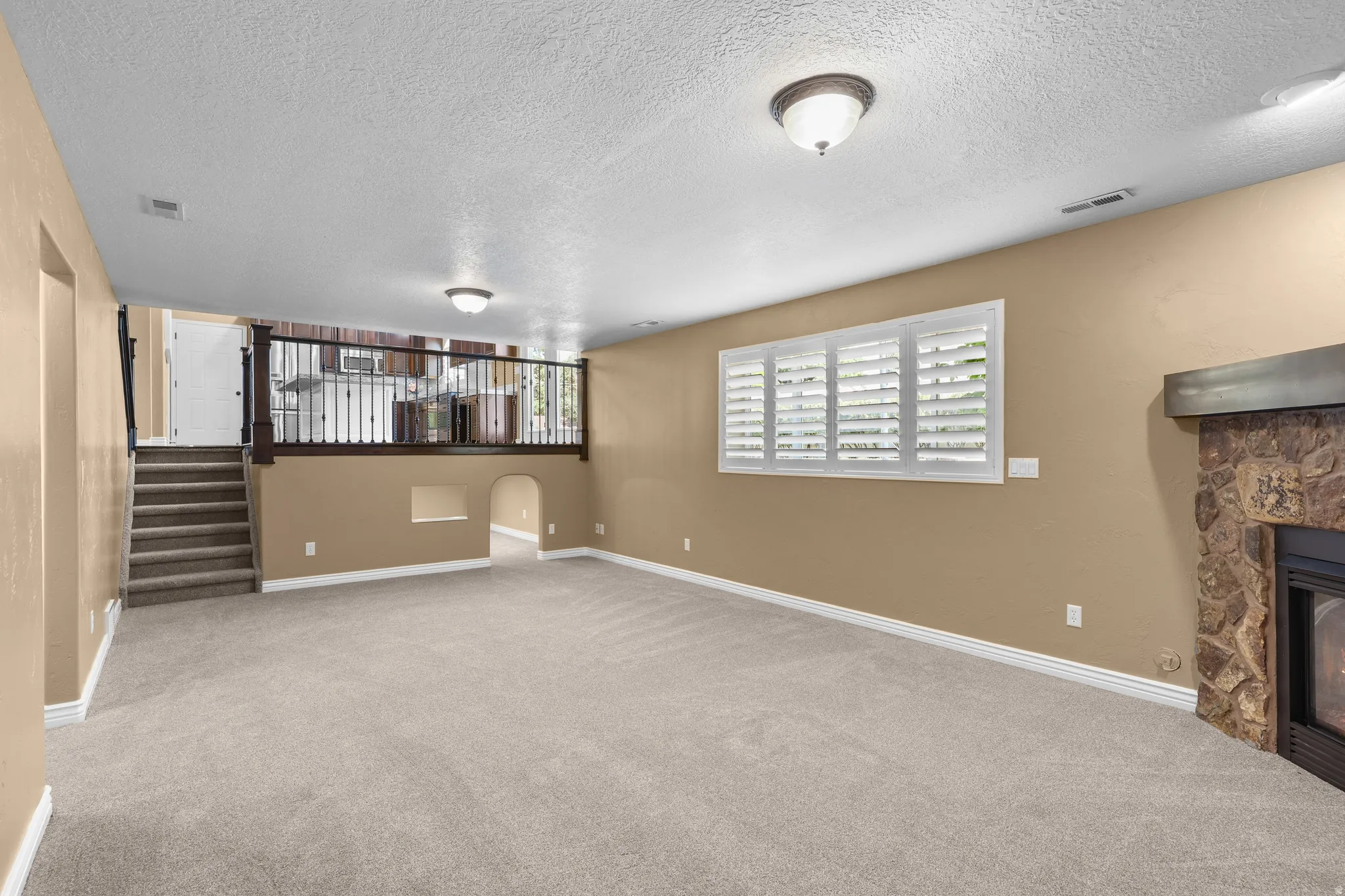 Carpeted lower-level room featuring a built-in stone fireplace, window with plantation shutters, staircase with wood railing, and arched pass-through