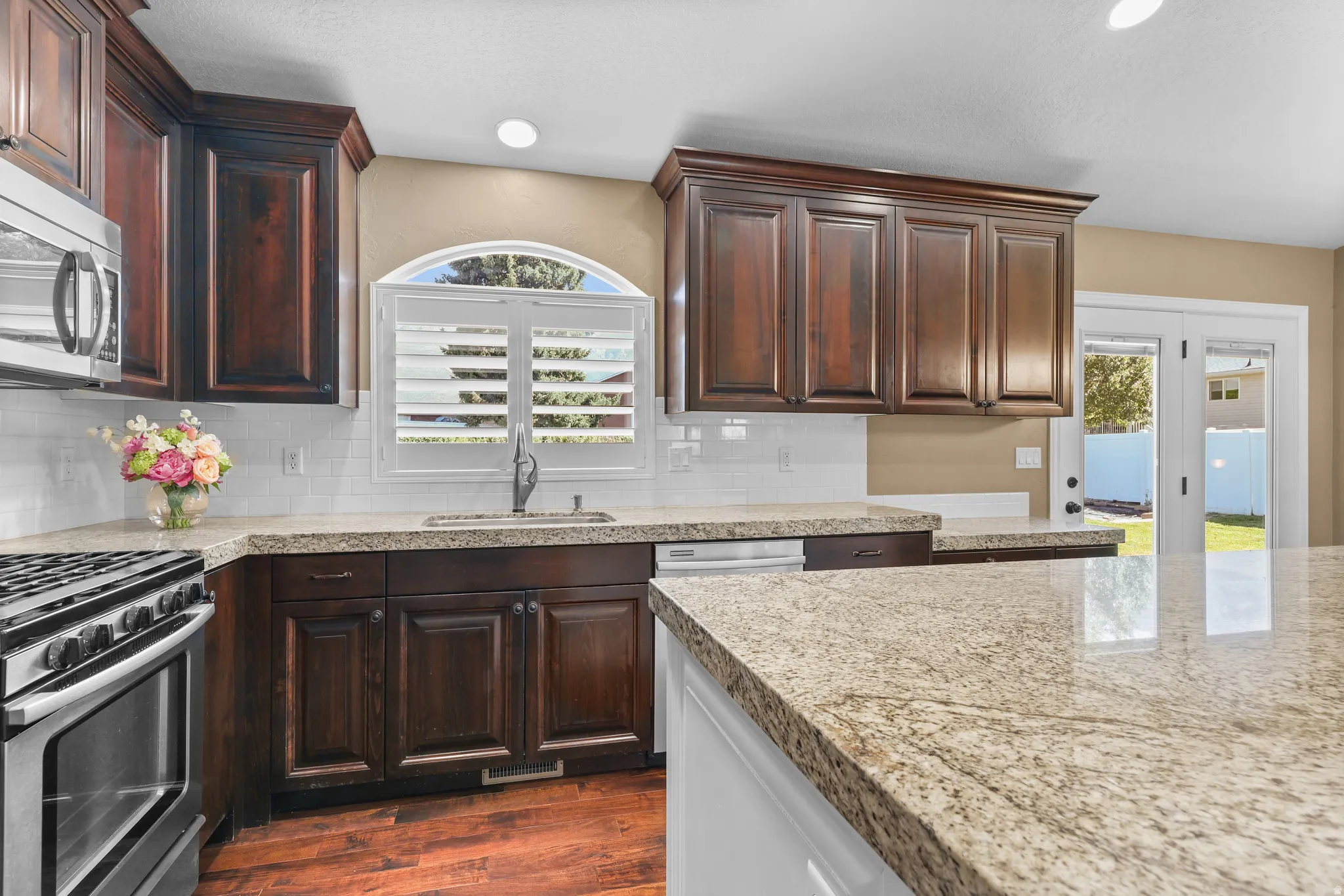 Kitchen featuring dark wood cabinetry, light granite countertops, stainless steel appliances, a subway tile backsplash, and wood-finish flooring