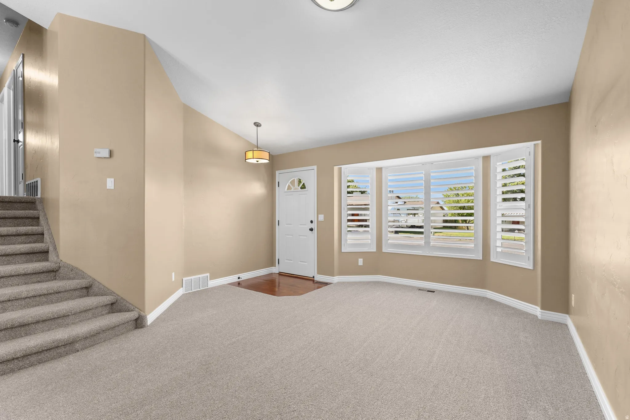 Carpeted living area featuring a bay window with plantation shutters, a white entry door with an arched window, and a wood-finish flooring entryway