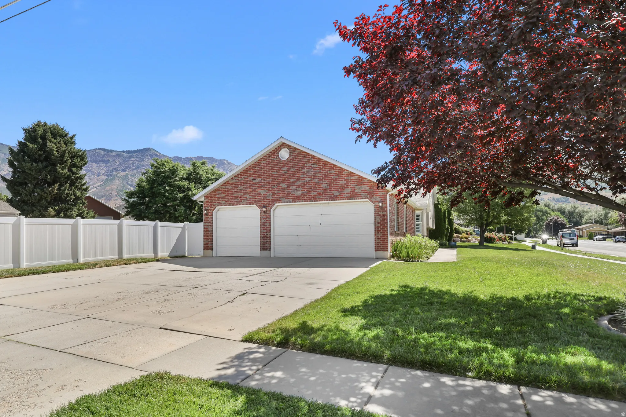 Brick exterior featuring a two-car garage with white doors, expansive concrete driveway, manicured lawn, and white vinyl fencing