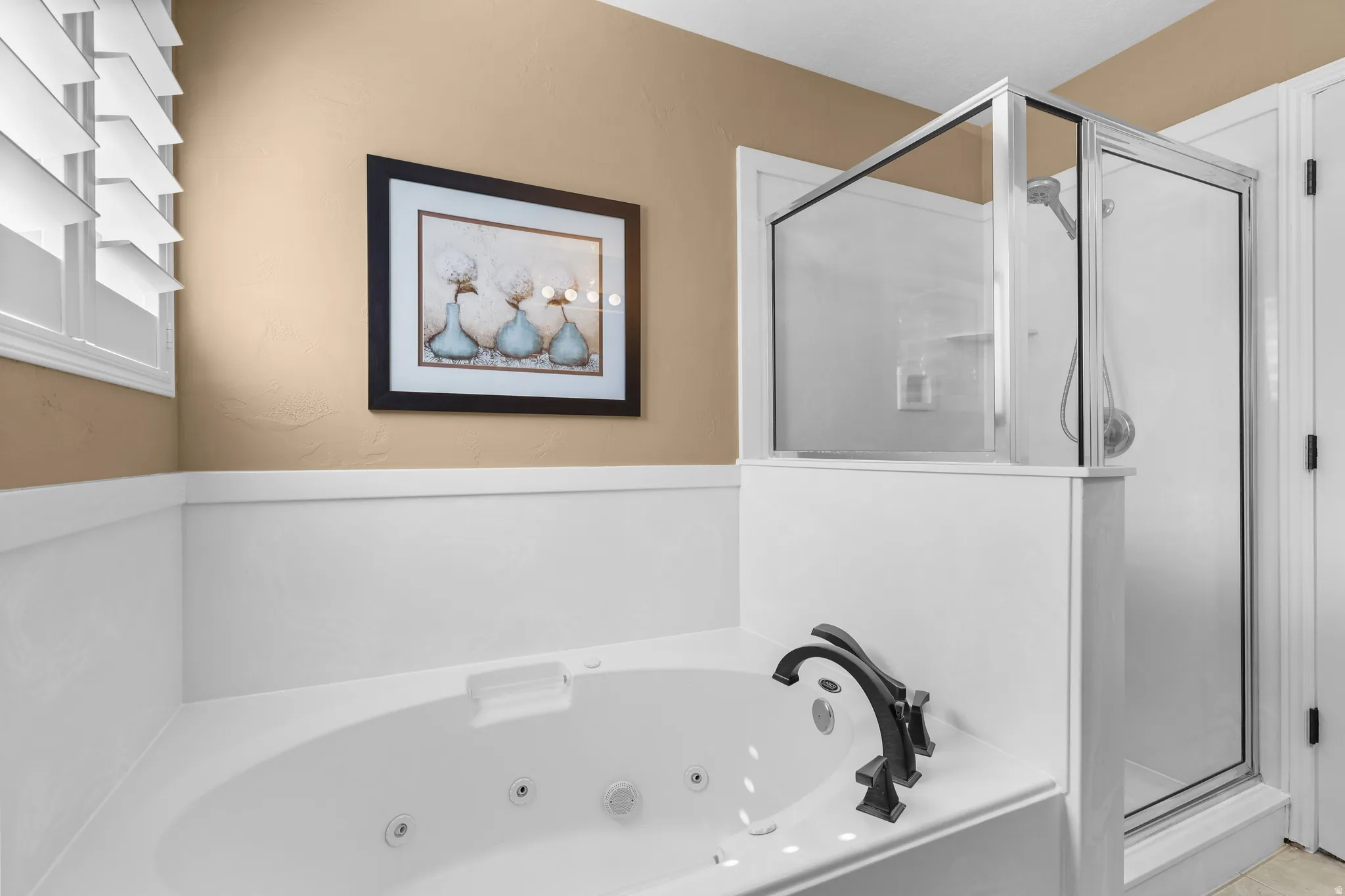 Bathroom featuring a built-in jetted tub with dark bronze fixtures, a glass-enclosed shower, plantation shutters, and wainscoting
