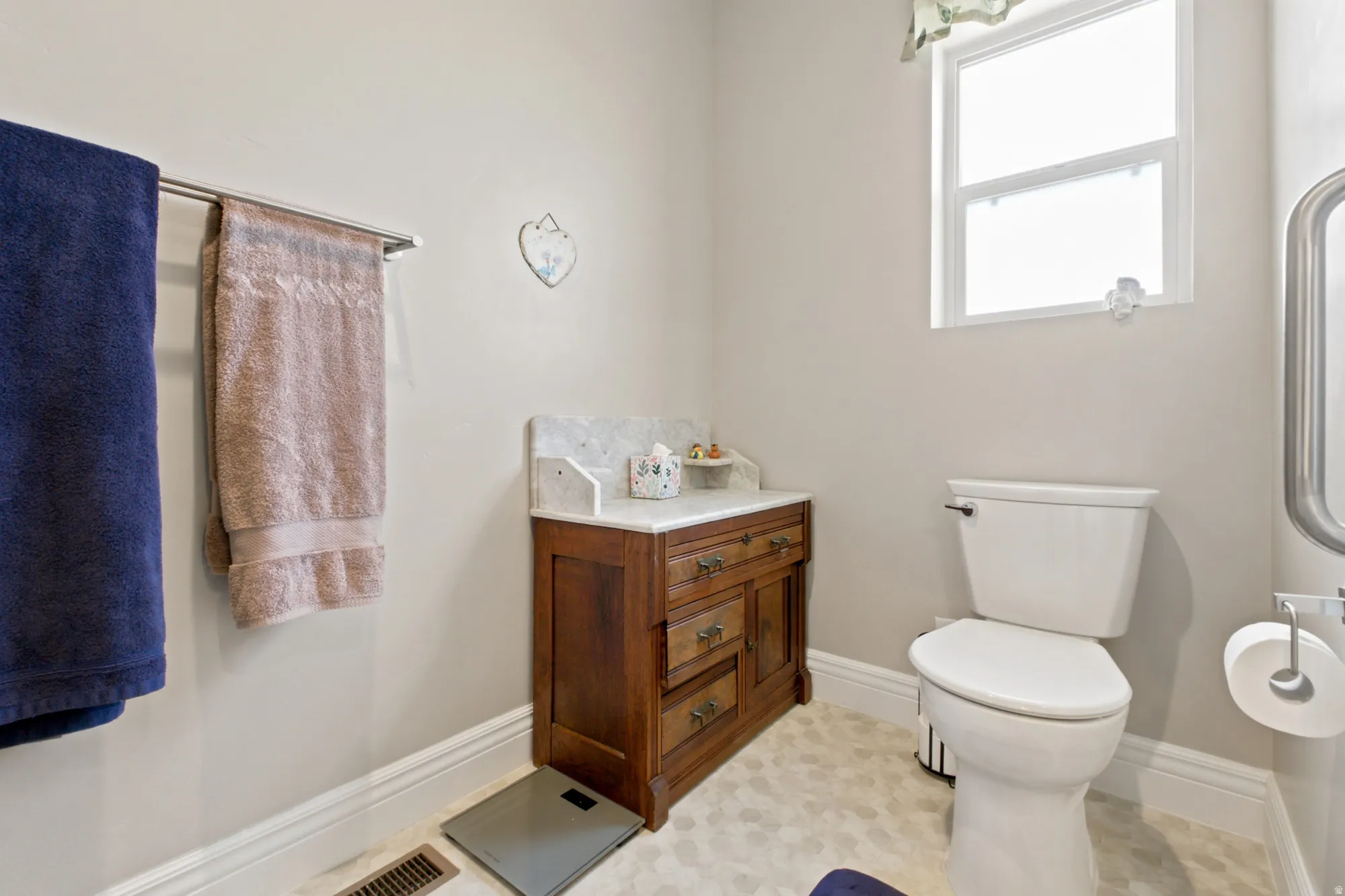 Bathroom featuring a white toilet, wood-finish vanity with a marble countertop, window with white trim, and light hexagonal tile flooring