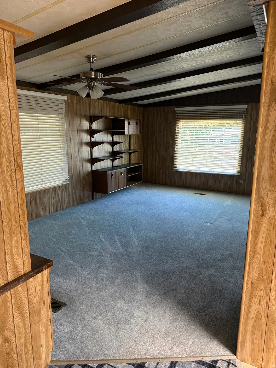 Spacious room featuring blue carpeting, wood-paneled walls, and a ceiling with exposed dark beams and a ceiling fan