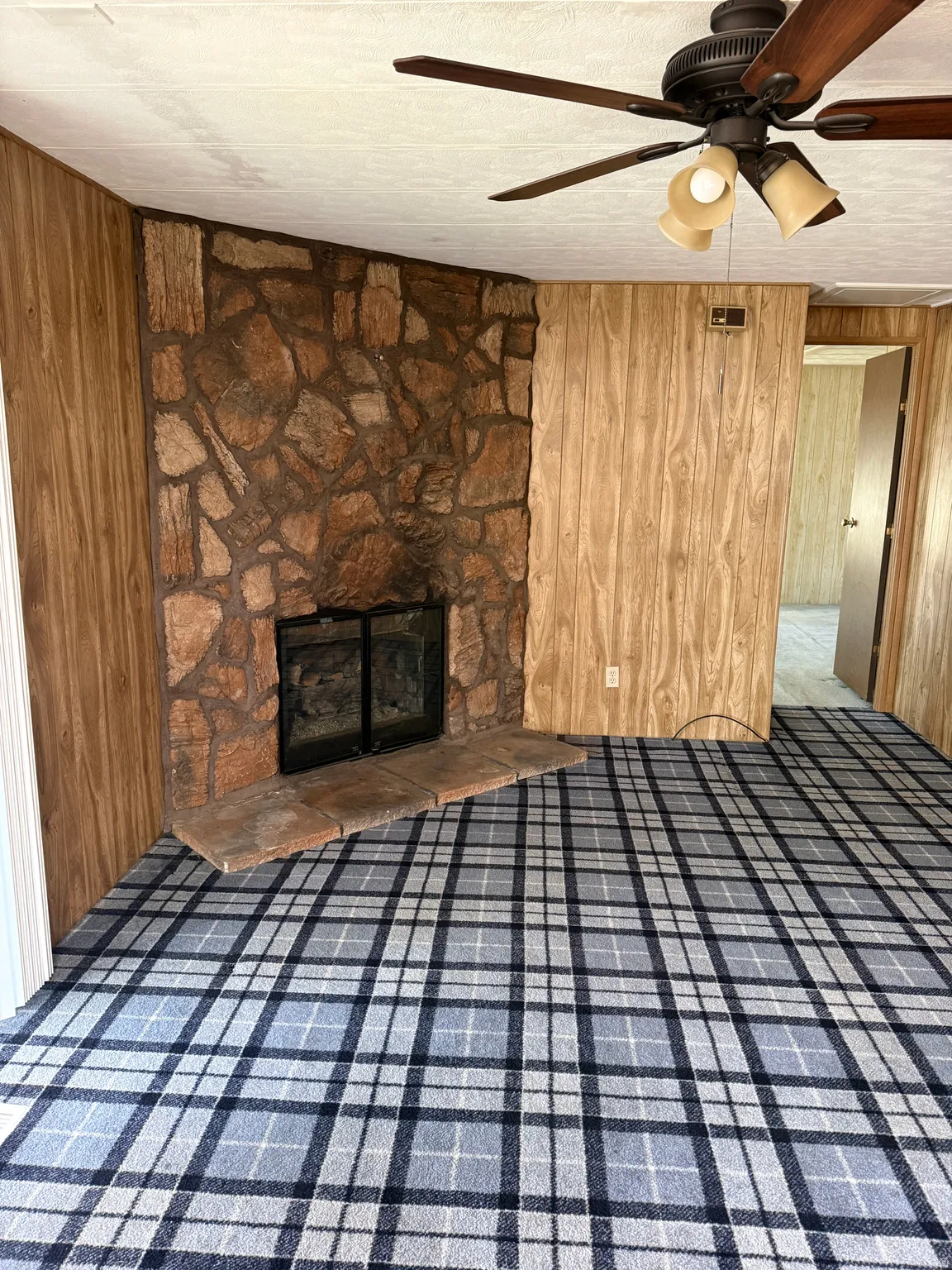 Stone fireplace with hearth, wood-finish paneling, patterned carpet, and ceiling fan with integrated lighting