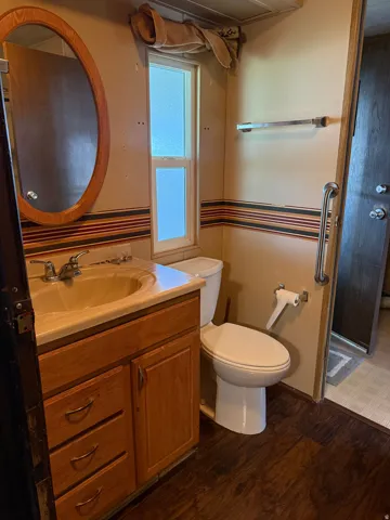 Bathroom featuring wood-finish flooring, a wood-finish vanity with an integrated sink, a wall-mounted mirror, a standard toilet, and a frosted glass window