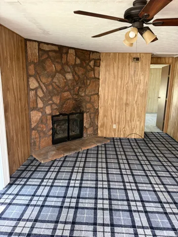 Stone fireplace with hearth, wood-finish paneling, patterned carpet, and ceiling fan with integrated lighting
