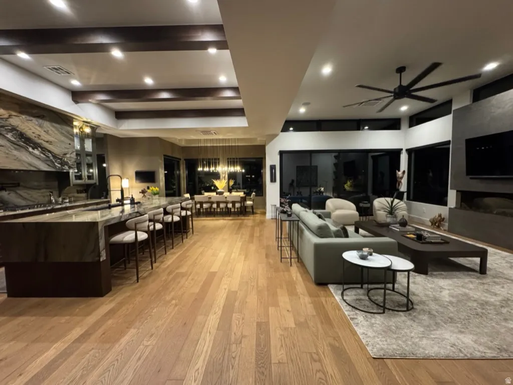 Open-concept living space featuring hardwood flooring, a large kitchen island with seating, a dining area with pendant lighting, a living area with a ceiling fan, and extensive window walls