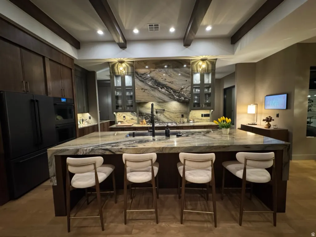 Gourmet kitchen featuring a waterfall island with integrated seating, stone countertops and backsplash, dark wood-finish cabinetry, and built-in black appliances