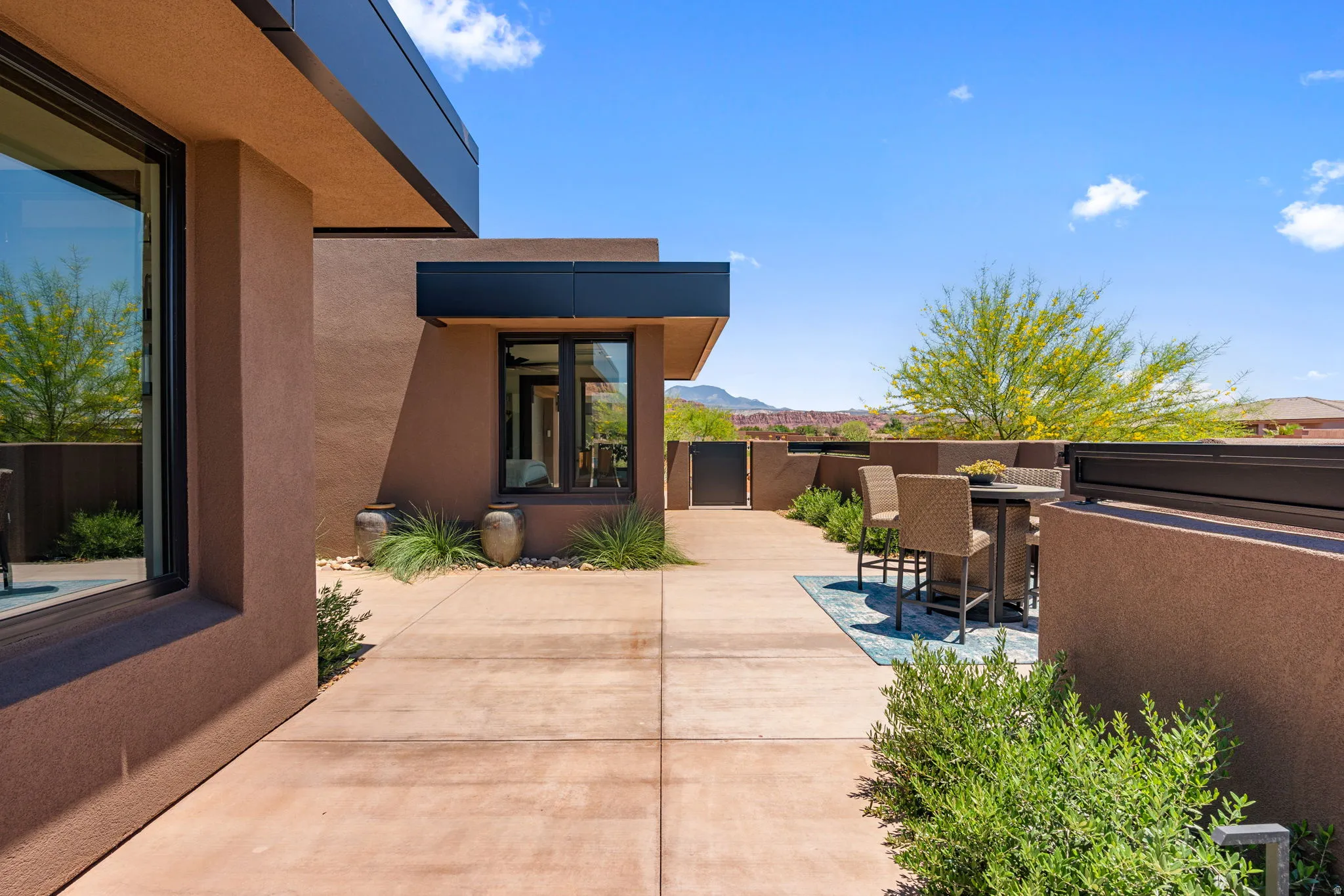 Exterior patio featuring expansive concrete paving, modern stucco finish, large dark-framed windows, and integrated landscaping with desert plants