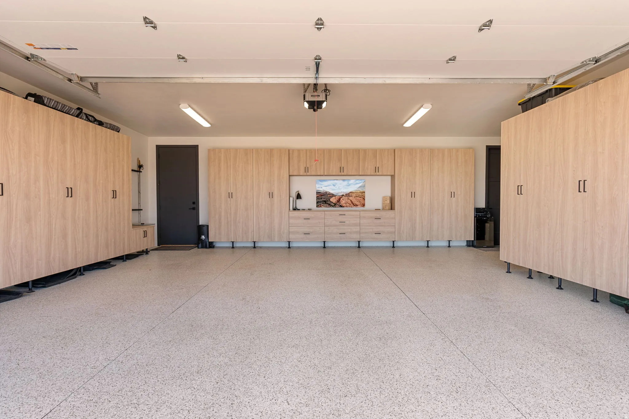 Epoxy-coated flooring in a spacious garage featuring extensive custom cabinetry, built-in storage, a dark entry door, and fluorescent overhead lighting