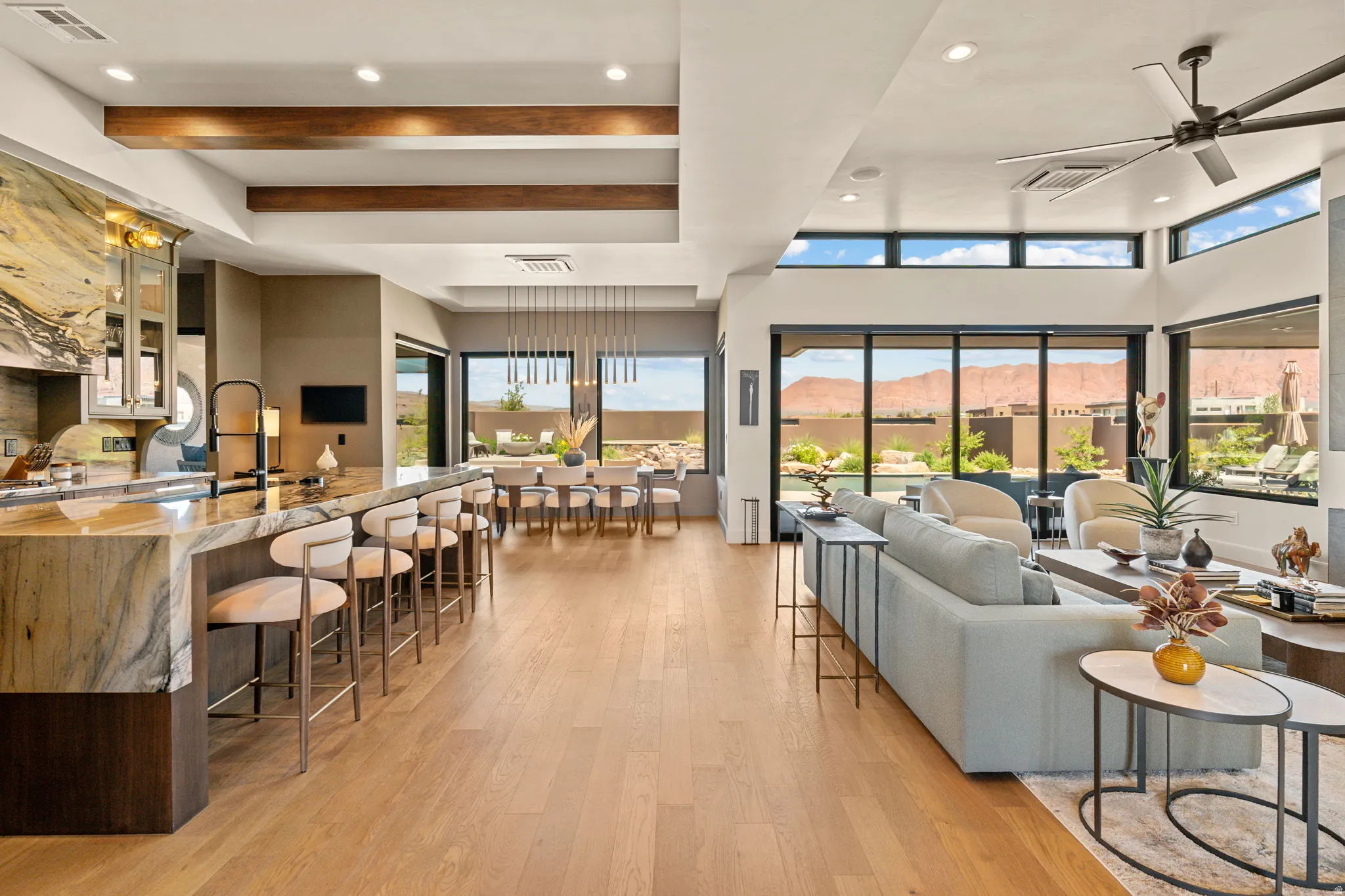 Open-concept living area featuring hardwood flooring, a kitchen island with stone countertops, a dining area with a linear chandelier, large glass sliding doors, and a ceiling fan