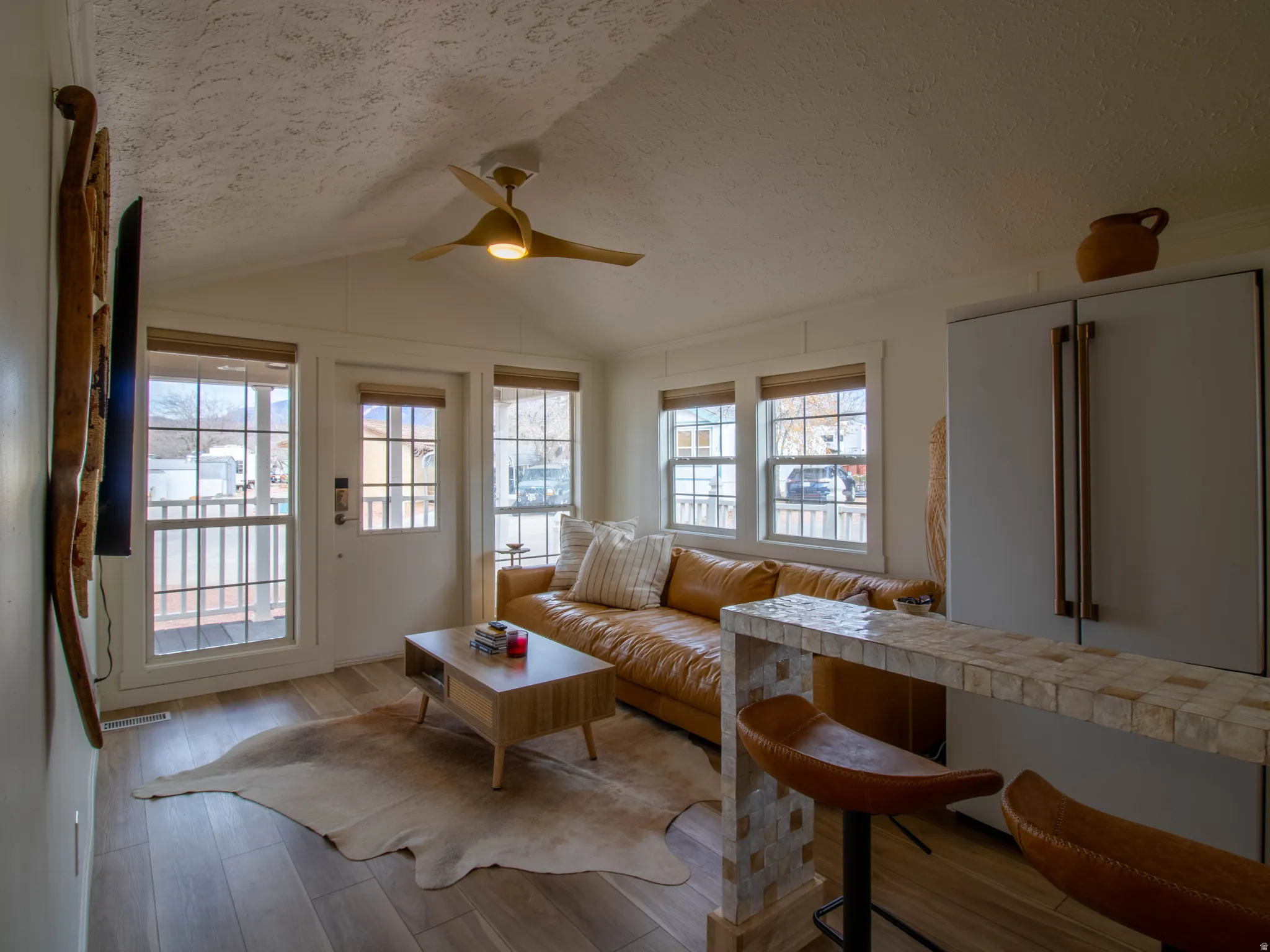 Living area featuring wood-finish flooring, a vaulted ceiling with a modern ceiling fan, multiple windows with blinds, a built-in breakfast bar with a patterned surface, and a full-size refrigerator with large handles