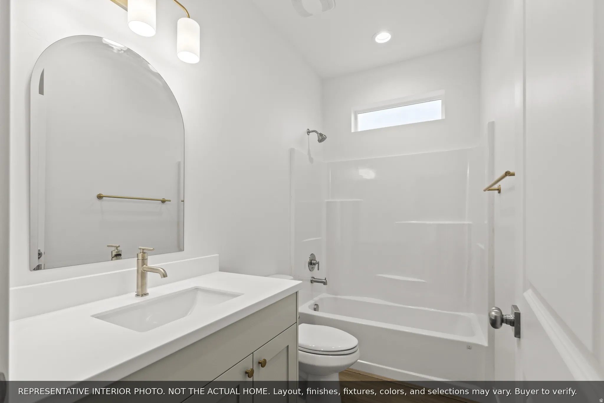 Bathroom featuring a white vanity with an integrated sink and brushed gold-tone faucet, an arched mirror, and a white tub/shower combination with a horizontal window