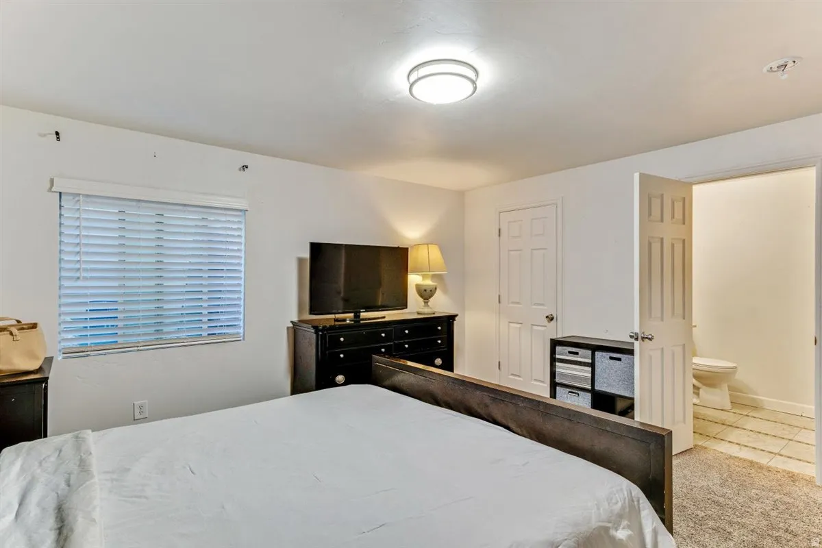 Bedroom with white walls, a flush-mount ceiling light, a window with horizontal blinds, and a paneled door opening to a tiled bathroom