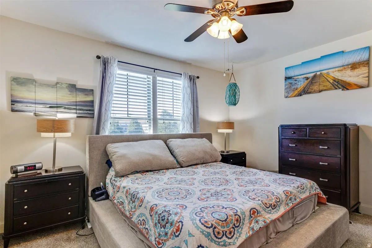 Carpeted bedroom featuring a large window with blinds and drapes, a ceiling fan with integrated lighting, and neutral wall tones
