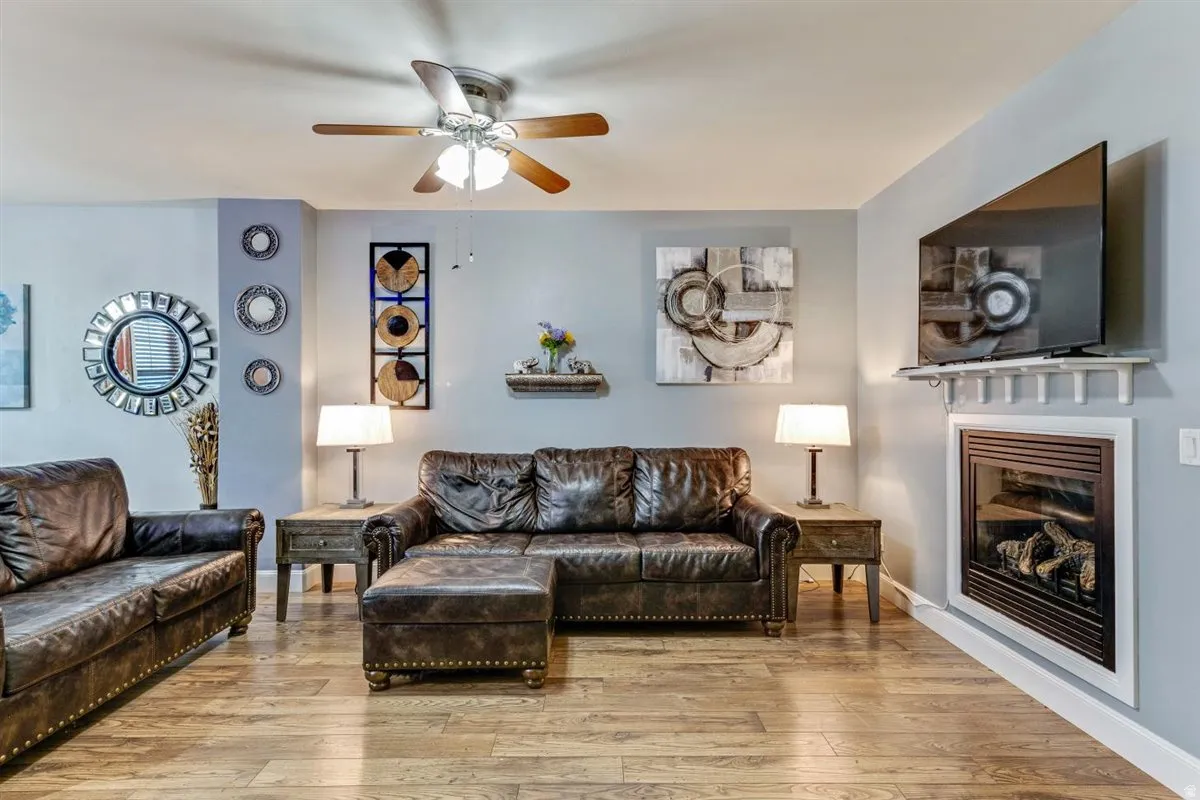 Living area featuring wood-finish flooring, a ceiling fan with integrated lighting, and a built-in fireplace with a white mantel shelf