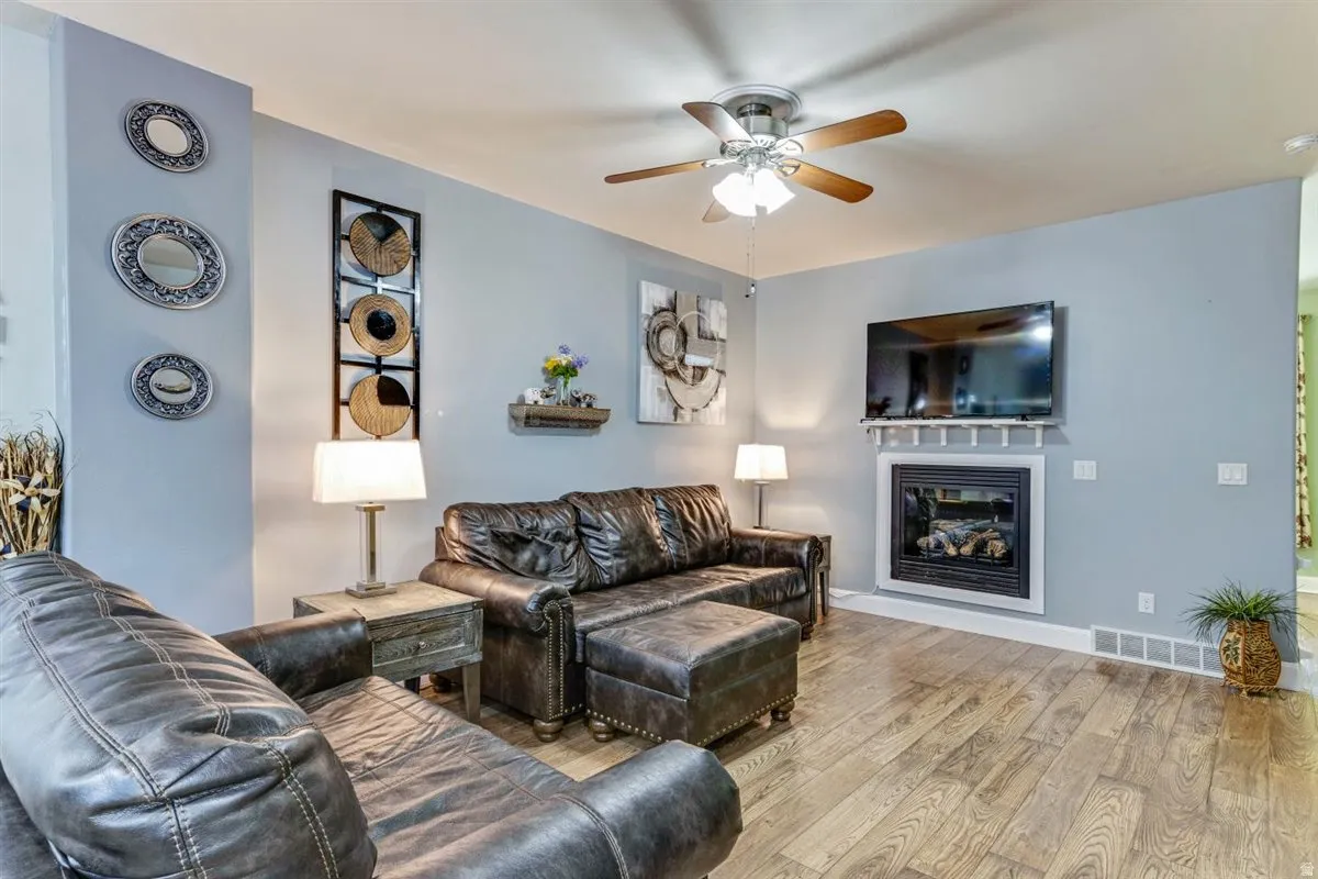 Living area featuring wood-finish flooring, a ceiling fan with integrated lighting, and a white-framed fireplace with a mantel
