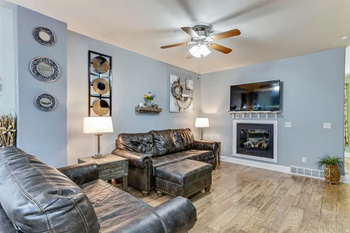 Living area featuring wood-finish flooring, a ceiling fan with integrated lighting, and a white-framed fireplace with a mantel