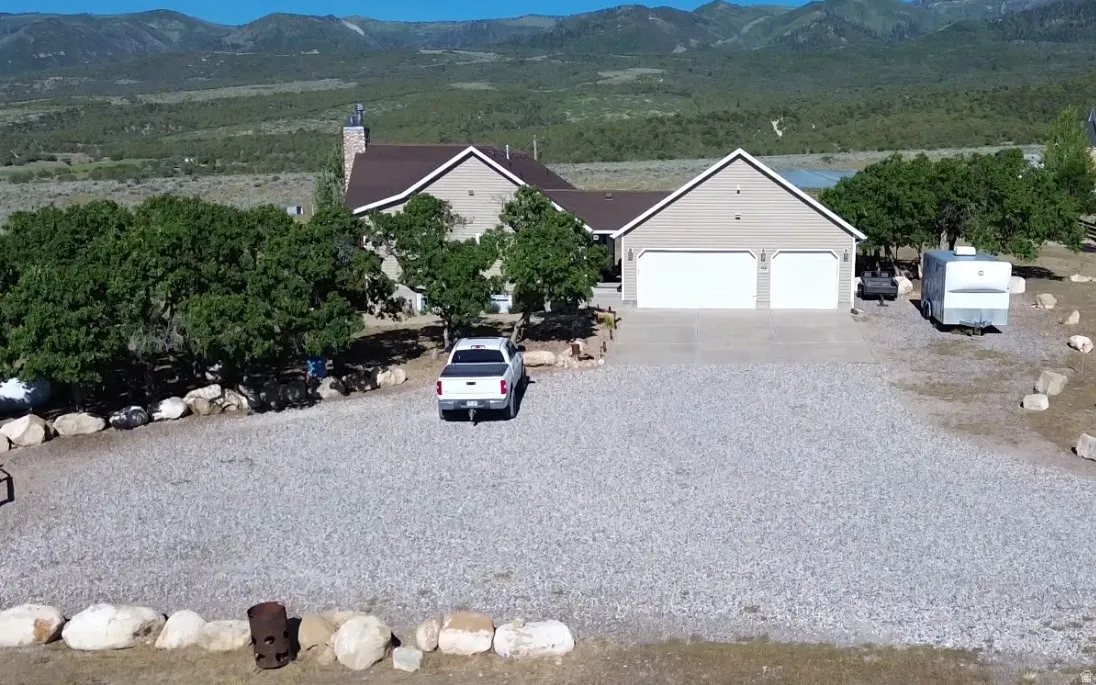 Mountain backdrop residence featuring a gravel driveway, mature trees, a two-car garage, and a stone-clad chimney