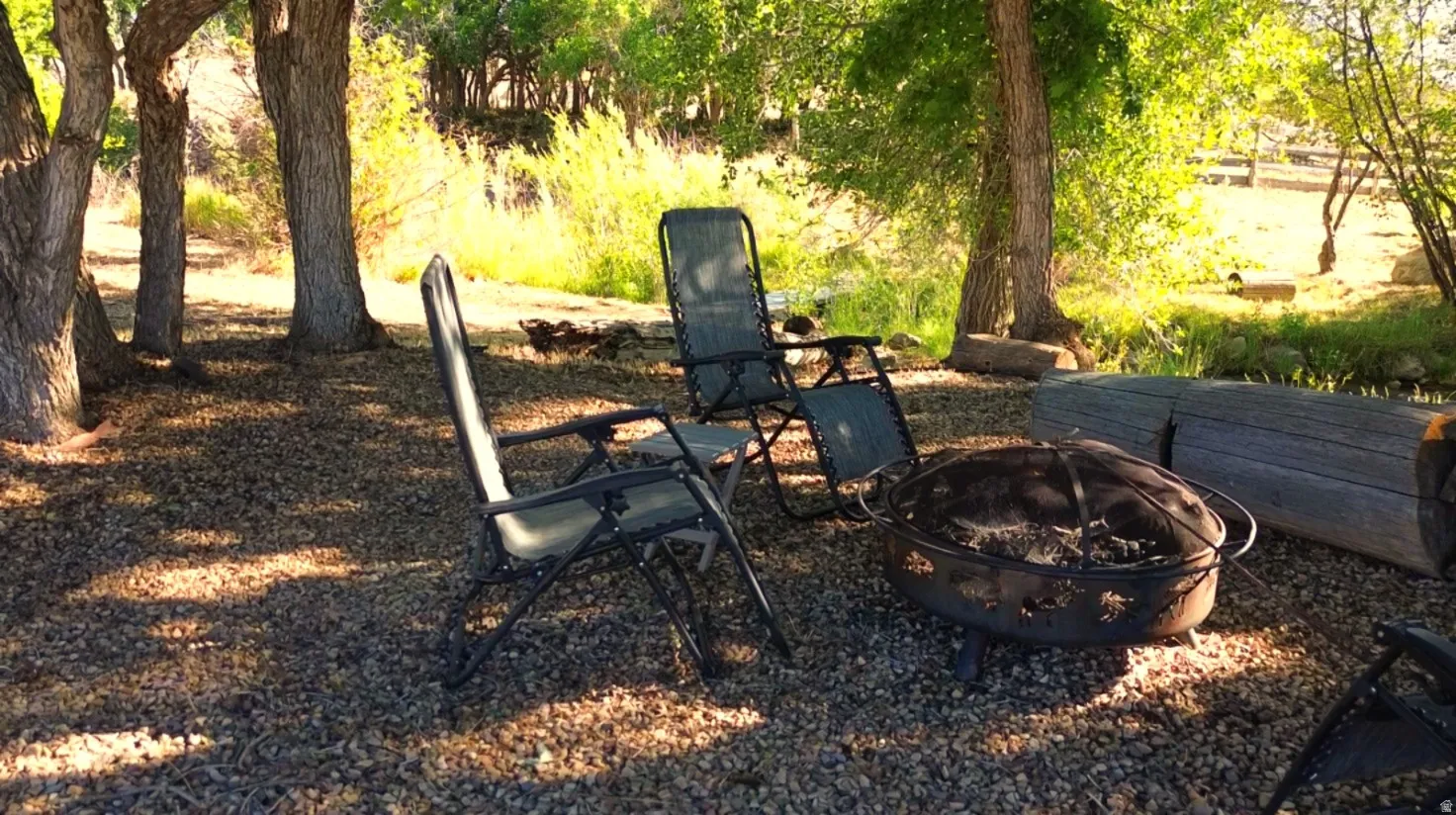 Outdoor gathering area featuring a gravel surface, mature trees providing shade, a fire pit, and natural wood log seating